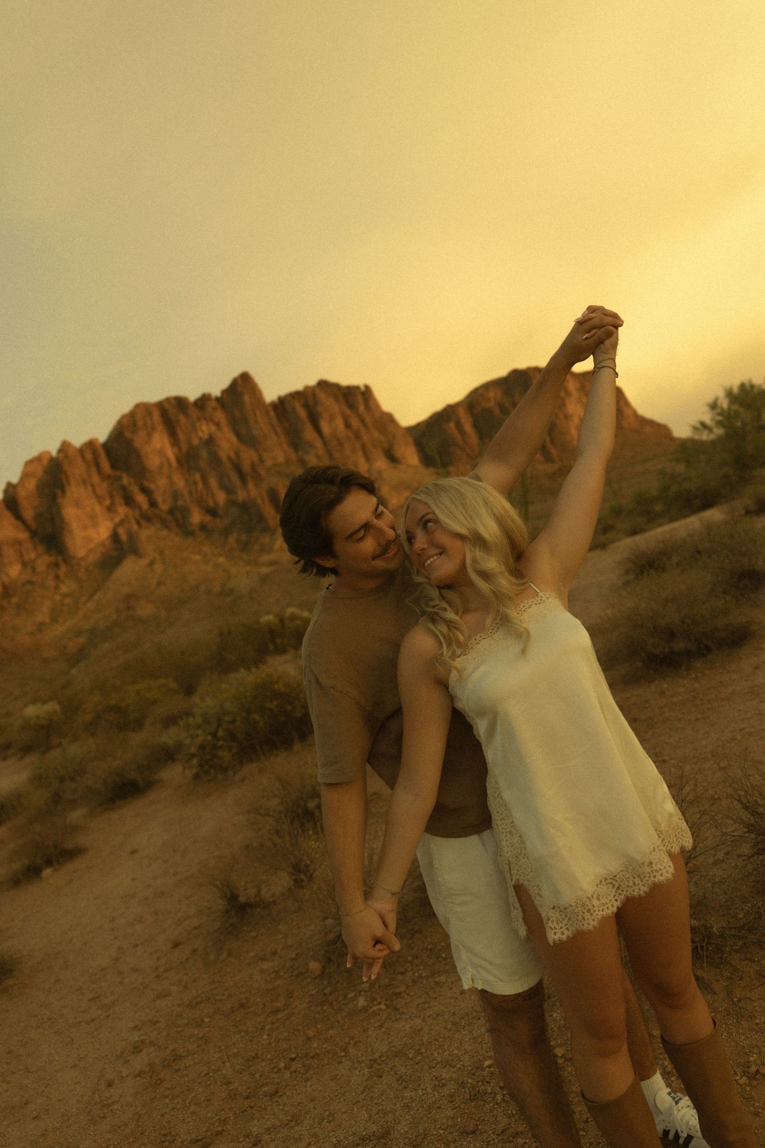 A joyful couple dances at sunset in a desert landscape with rocky mountains, the woman raising their joined hands.