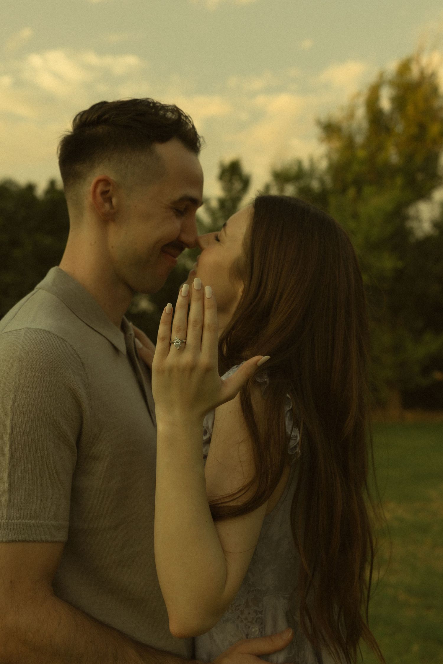 Couple sharing a romantic moment outdoors, woman showing engagement ring with green stone, trees in background.