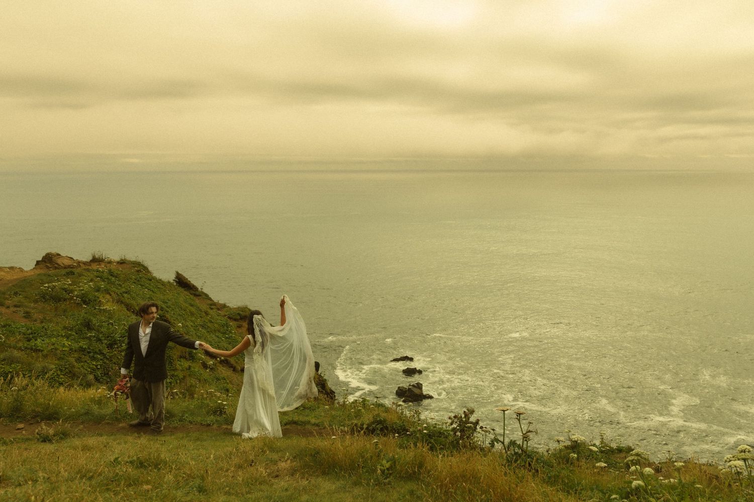 Bride and groom holding hands on a coastal cliff with ocean views and dramatic cloudy sky on their wedding day.