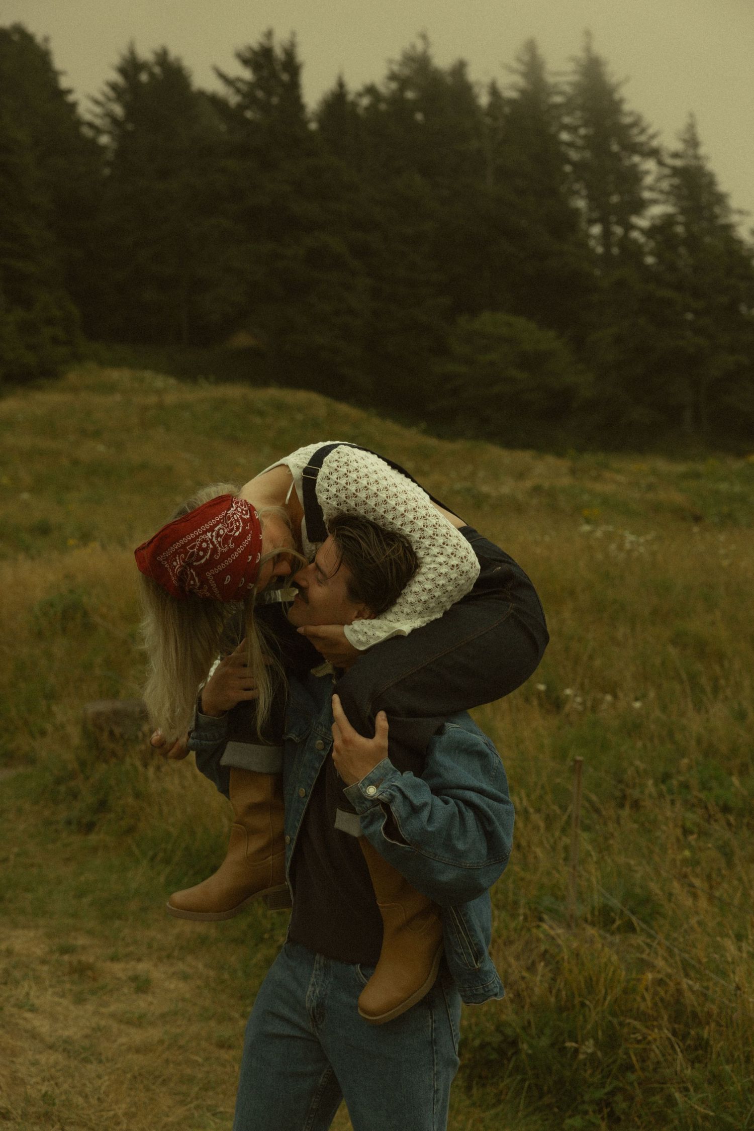 Couple sharing a romantic kiss in a grassy meadow with evergreen trees, woman wearing red bandana and boots.