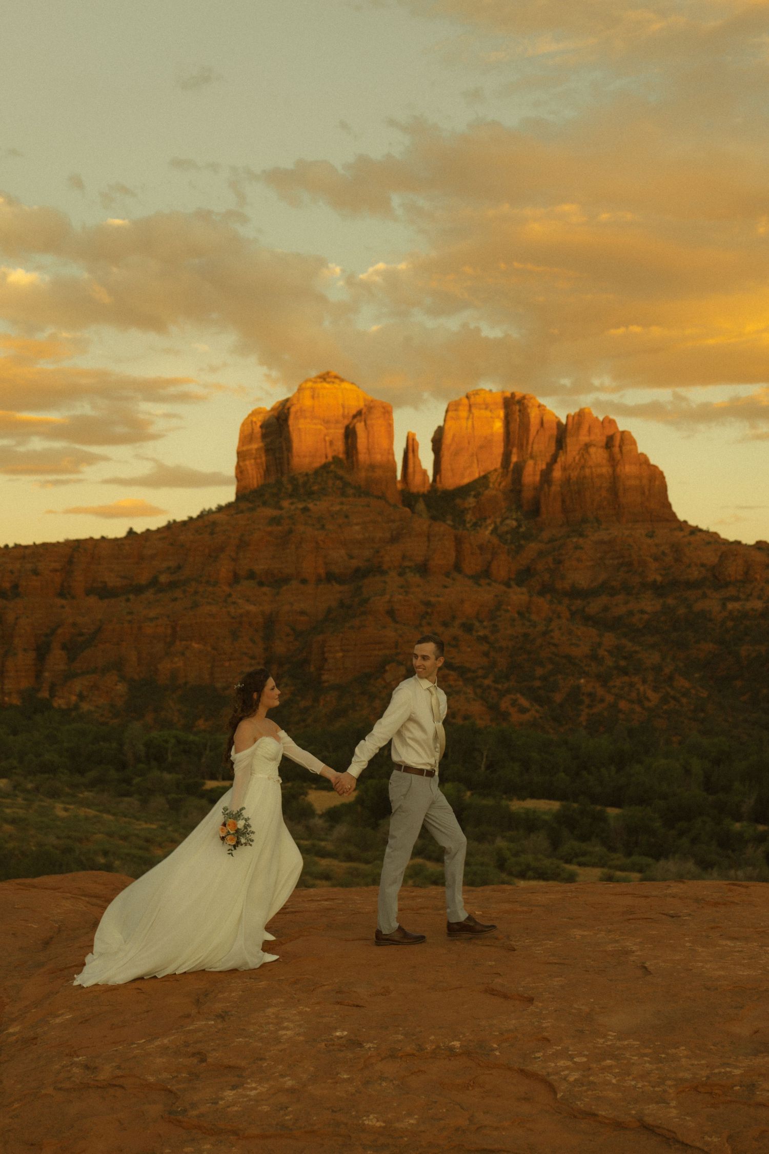Bride and groom hold hands at golden sunset with Cathedral Rock formation in Sedona, Arizona as backdrop.