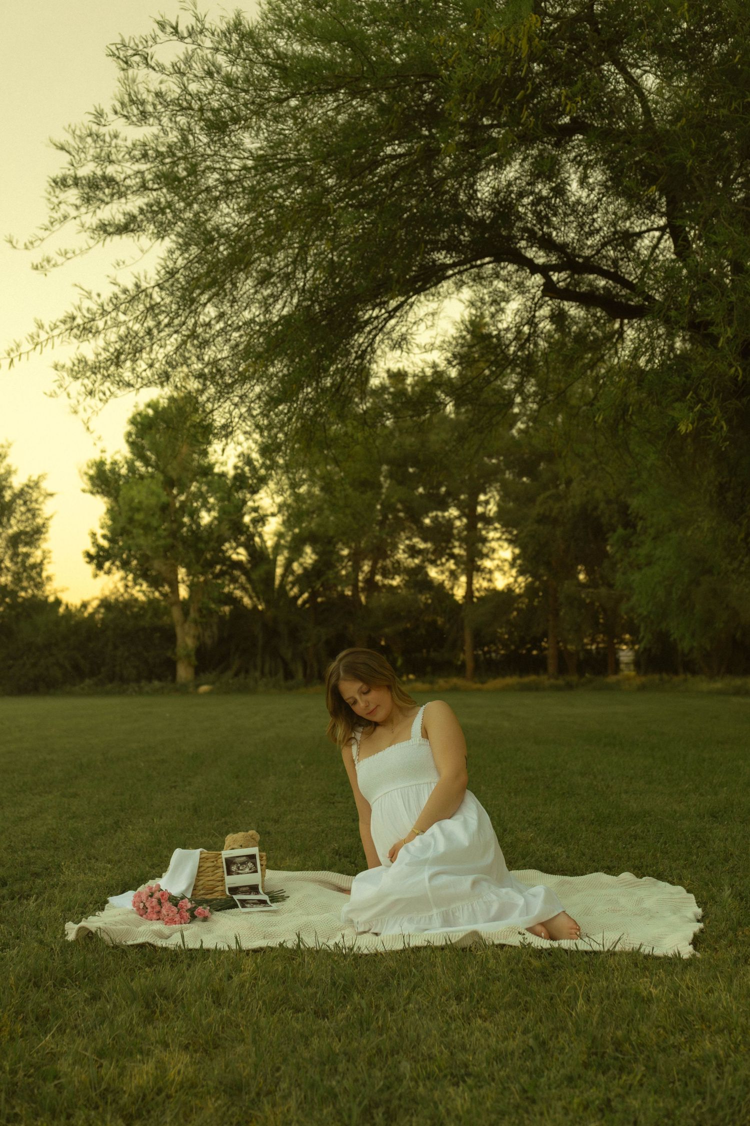 Pregnant woman in white dress sitting on blanket in park at sunset with ultrasound photos and flowers.