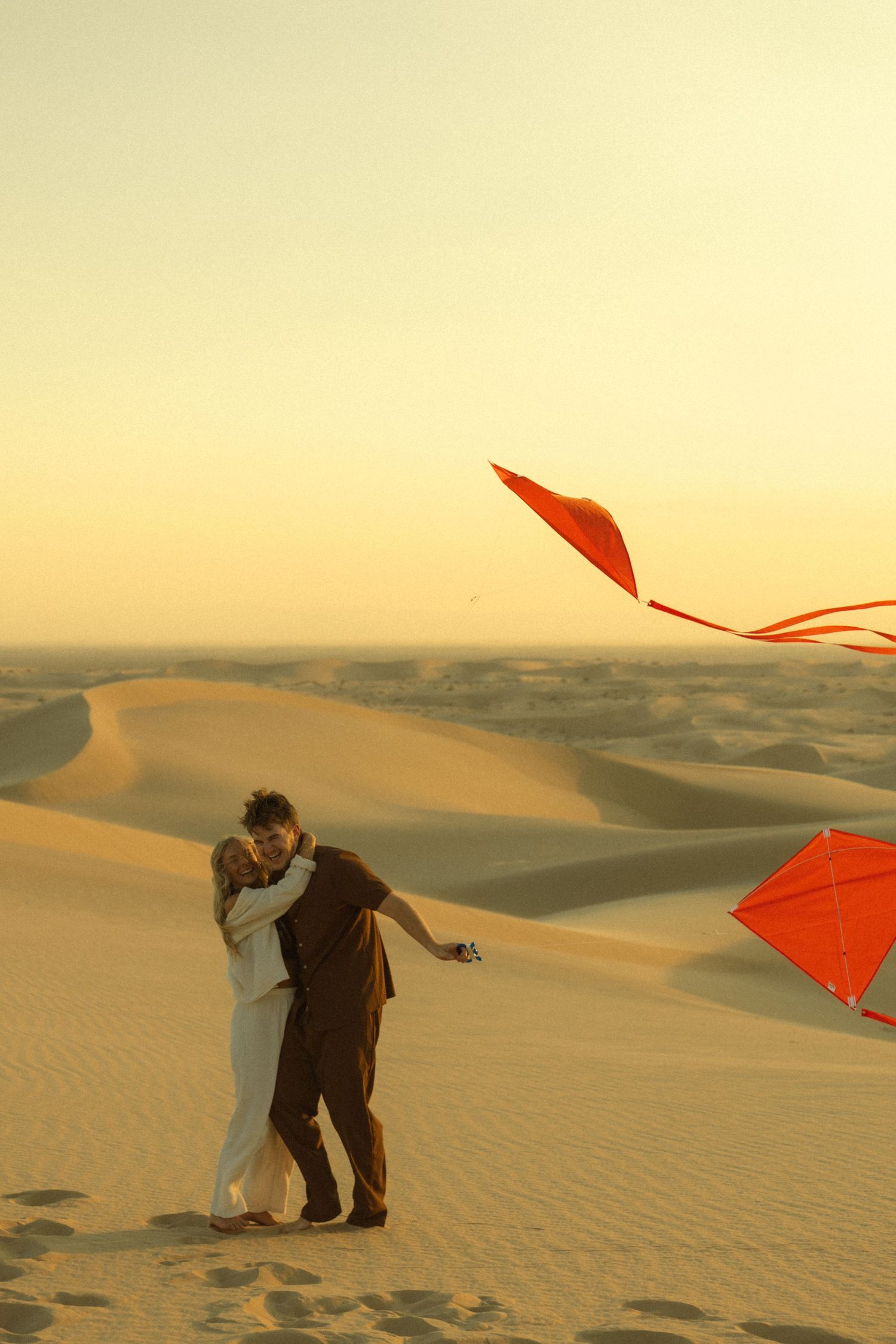 Couple embracing romantically on golden sand dunes at sunset while red kites soar above in desert landscape.