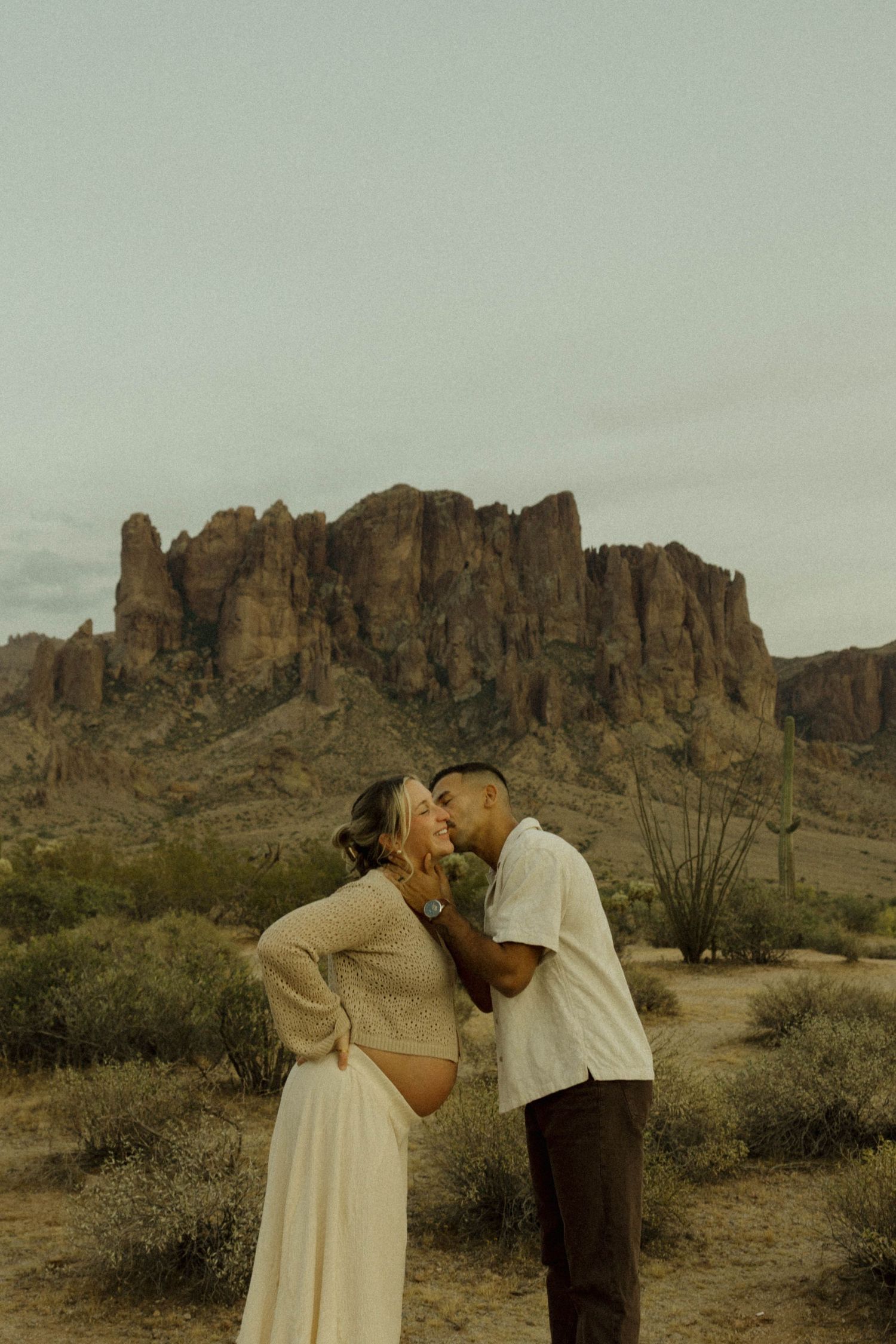 Couple sharing a kiss during maternity photo shoot in Arizona desert with Superstition Mountains backdrop.