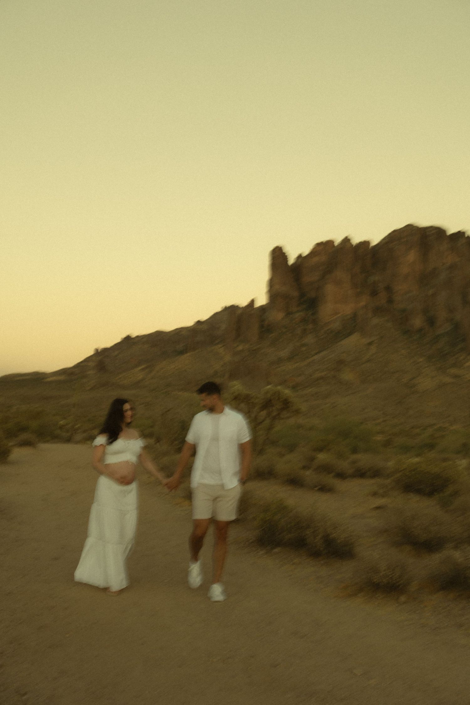 Couple in white outfits holding hands walking on desert path at sunset with rocky mountain backdrop.