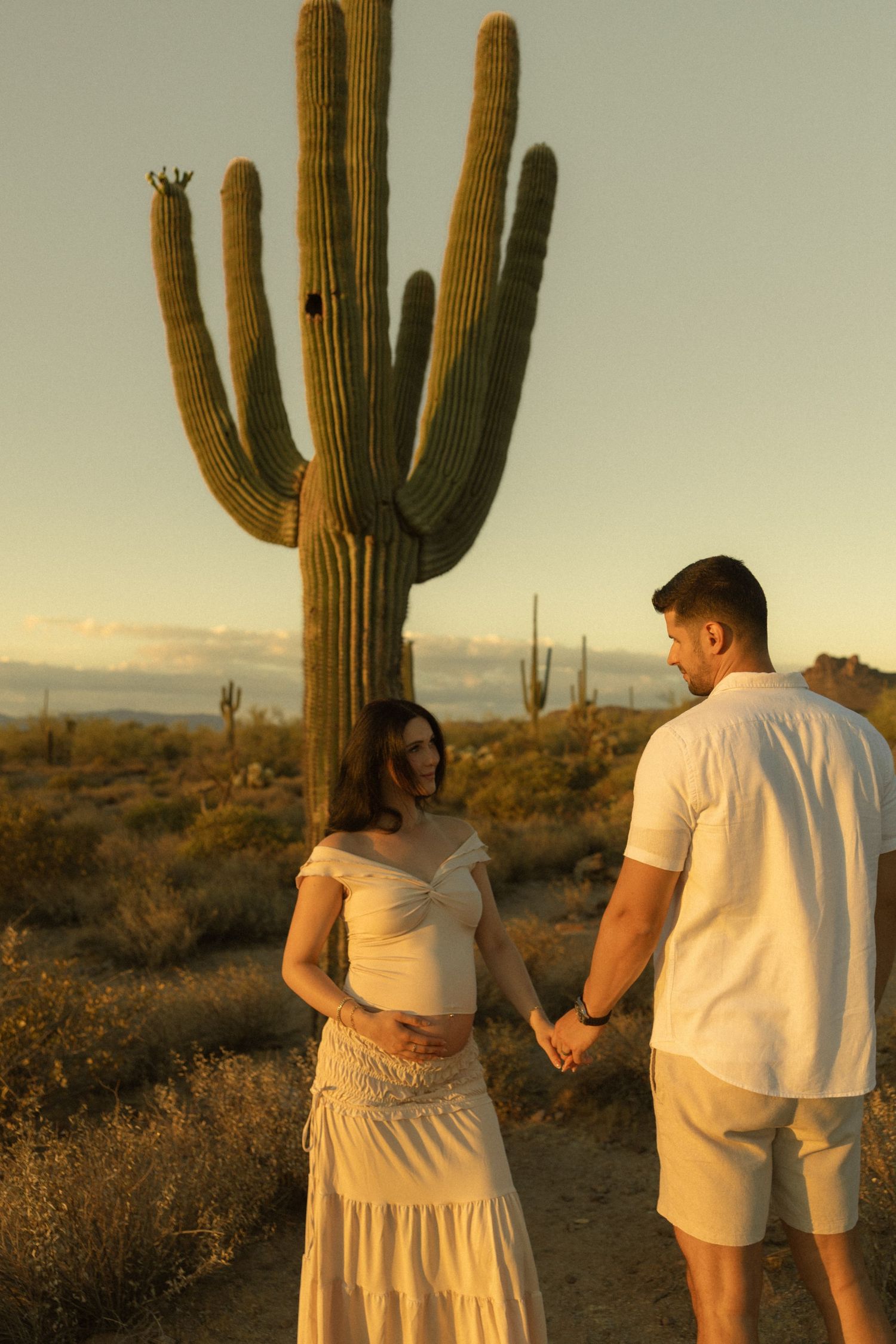 Expecting couple holding hands gazing at each other in desert landscape with saguaro cactus at dusk.