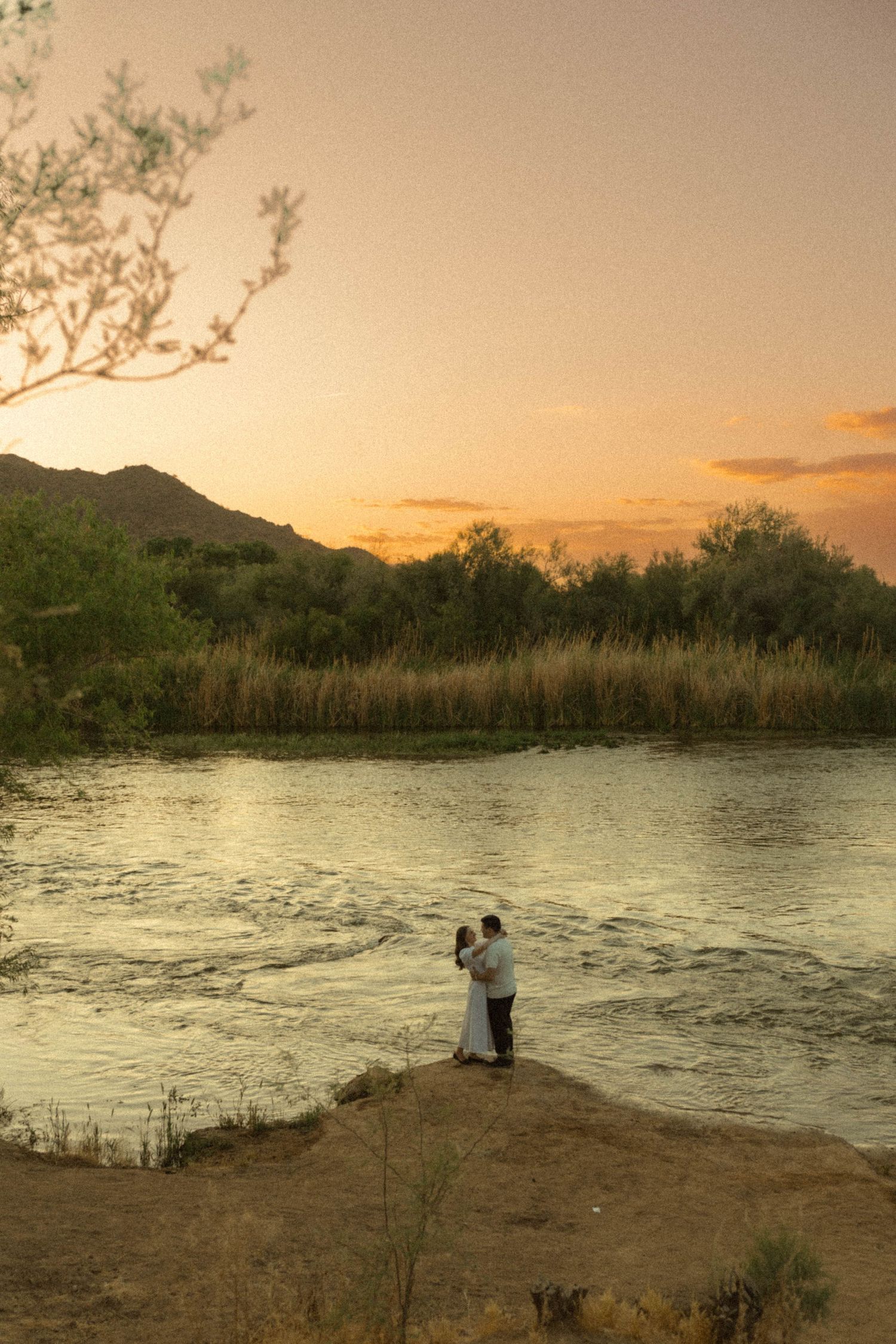 Couple embracing on a rocky riverbank at vivid orange sunset with mountains and reeds in background.