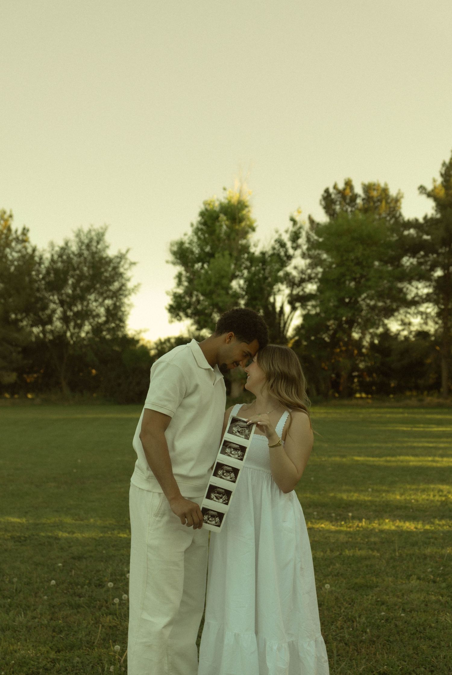 Couple in white outfits sharing a tender moment in a park, holding ultrasound photos to announce pregnancy.