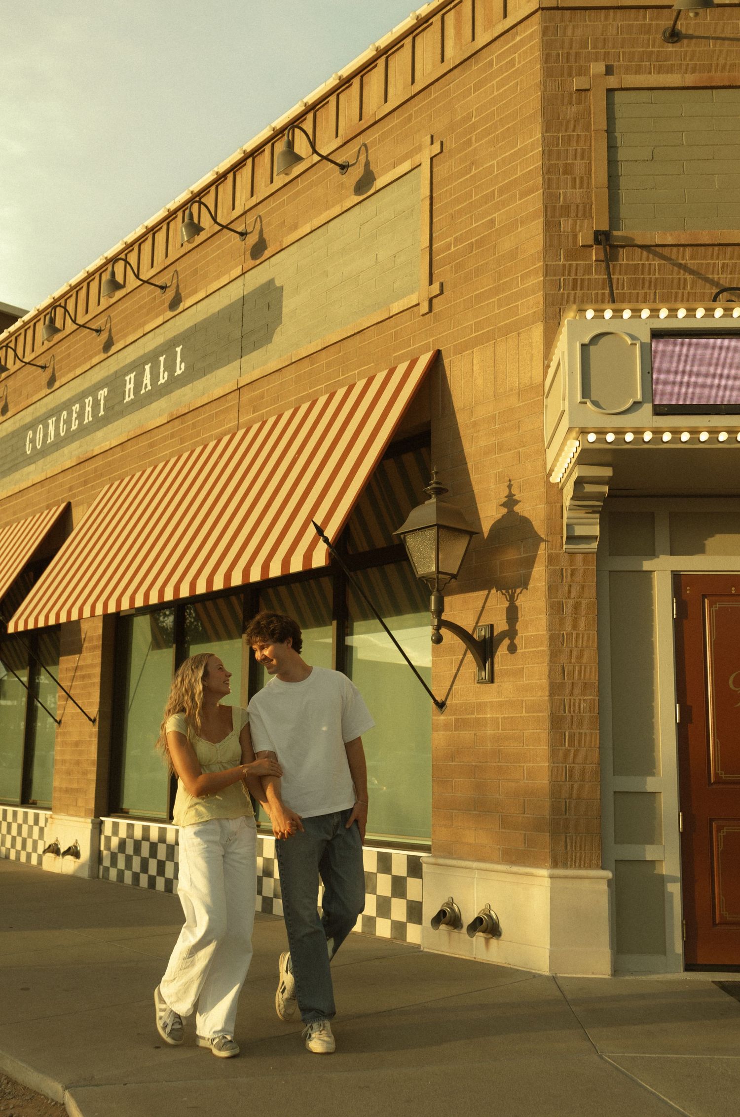 A couple strolls arm in arm outside a concert hall building with orange striped awnings at golden hour.