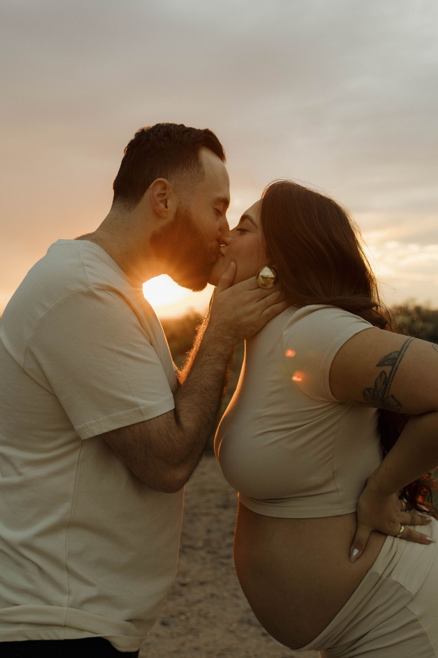 A romantic maternity photo at sunset shows a couple sharing a tender kiss while highlighting the pregnant belly.