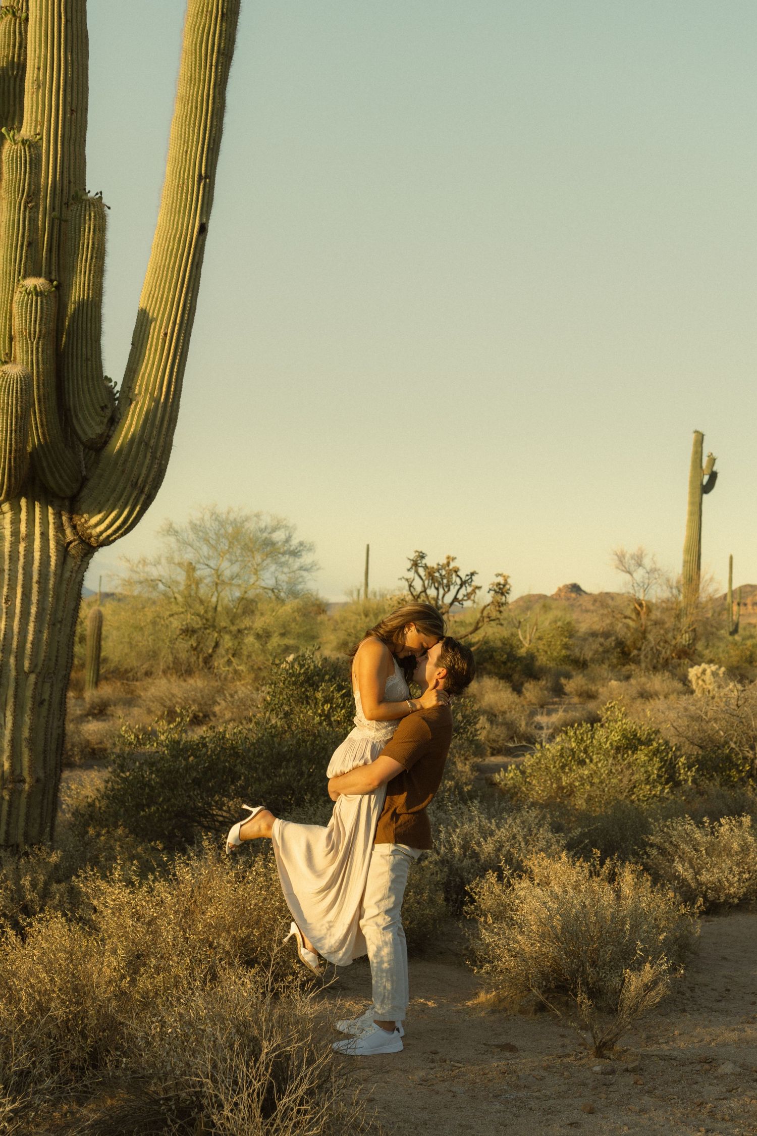 Couple sharing a romantic kiss at sunset in the Sonoran Desert, surrounded by tall saguaro cacti in Arizona.