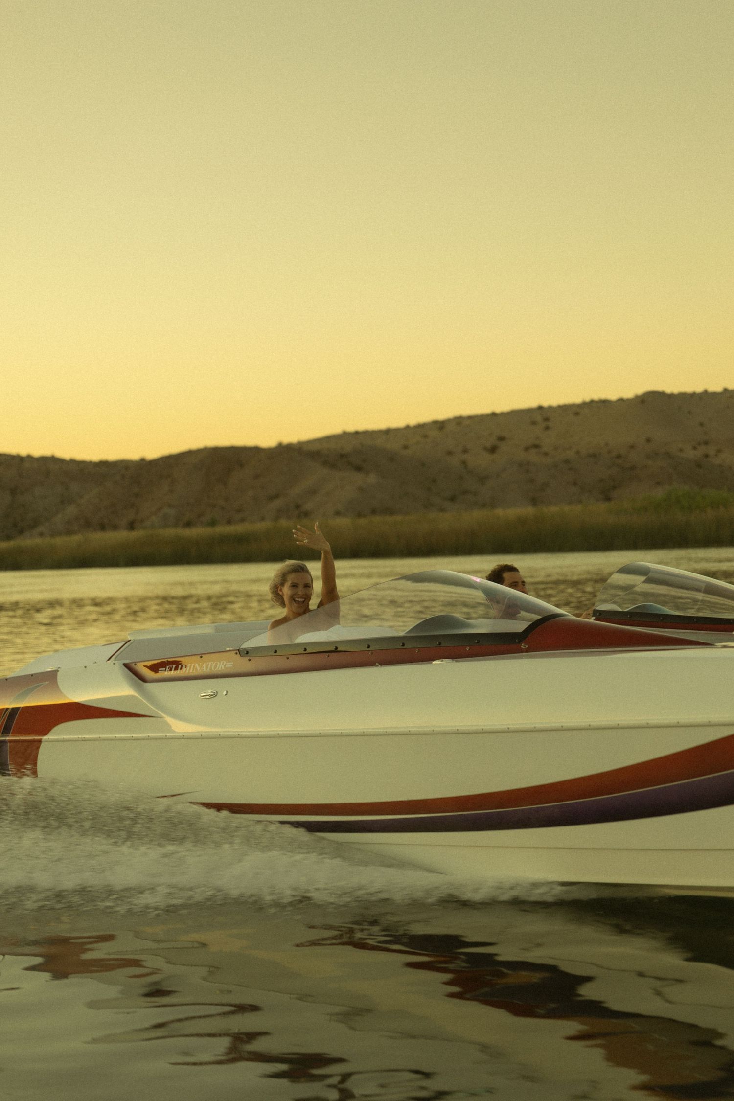 Woman waving from a speeding white and red motorboat on a lake during a warm golden hour sunset.