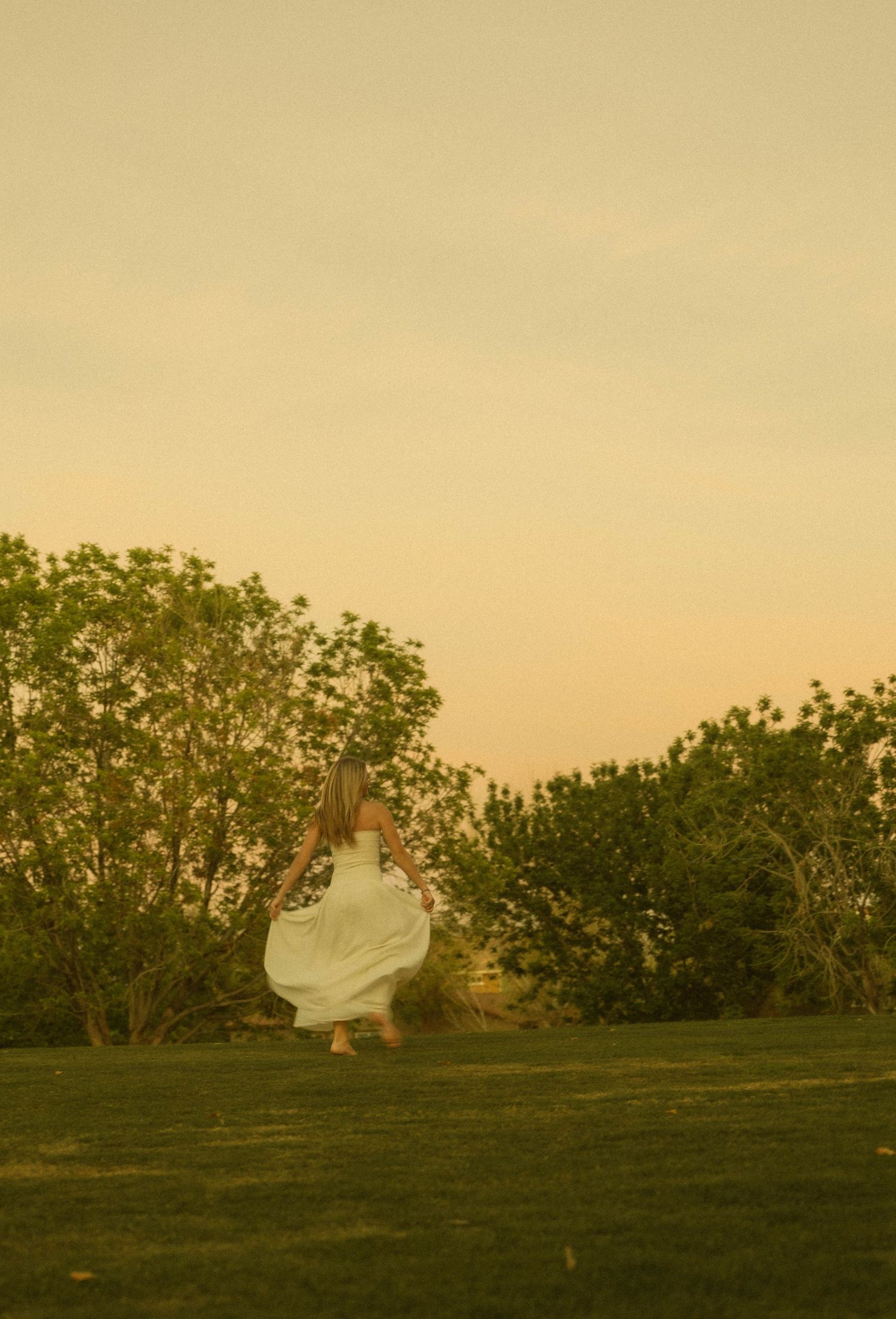 Woman in flowing white dress twirling near trees during a warm golden sunset.