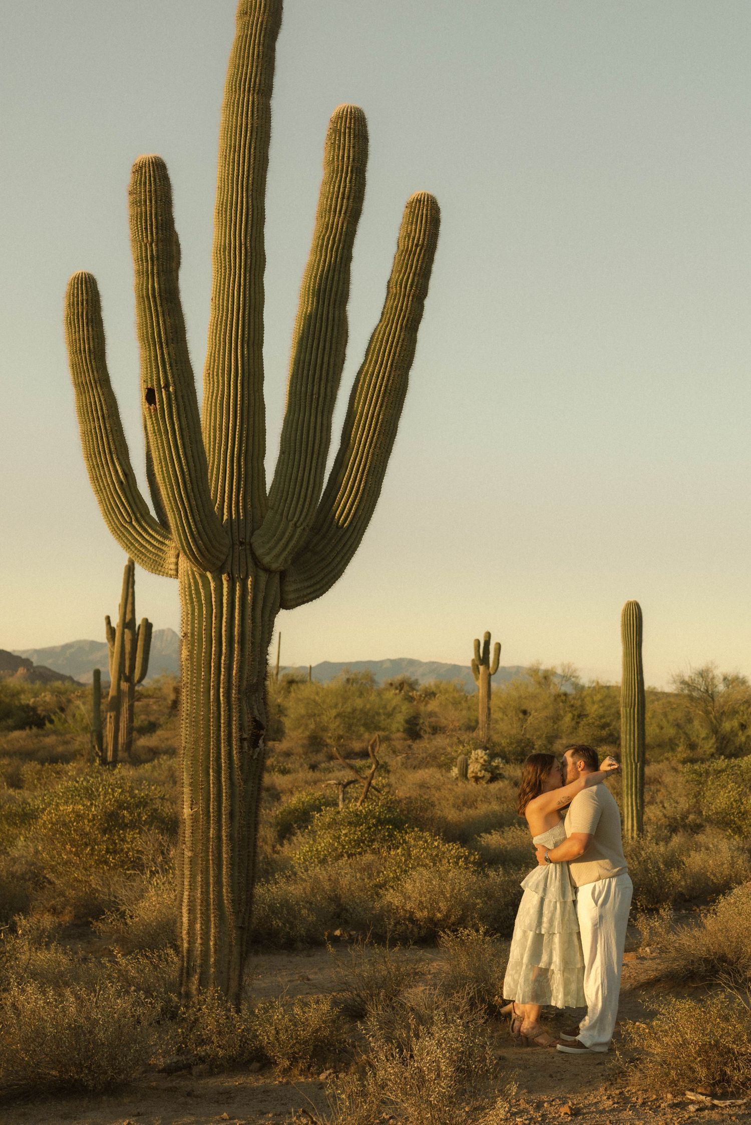 Couple embracing in white attire beside a tall saguaro cactus in the Arizona desert at golden hour.