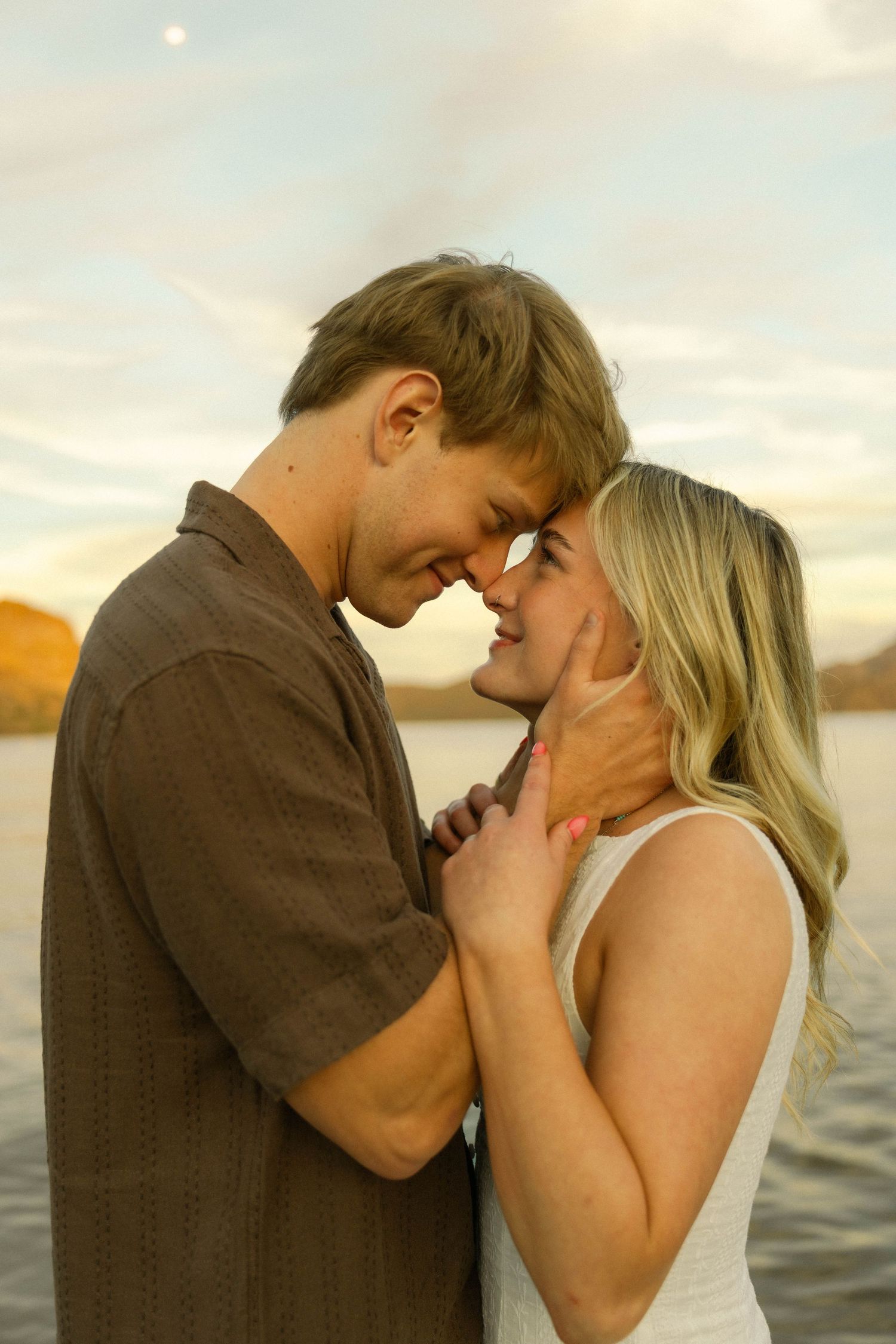 A couple embraces intimately on a dock during golden hour with a scenic mountain landscape.