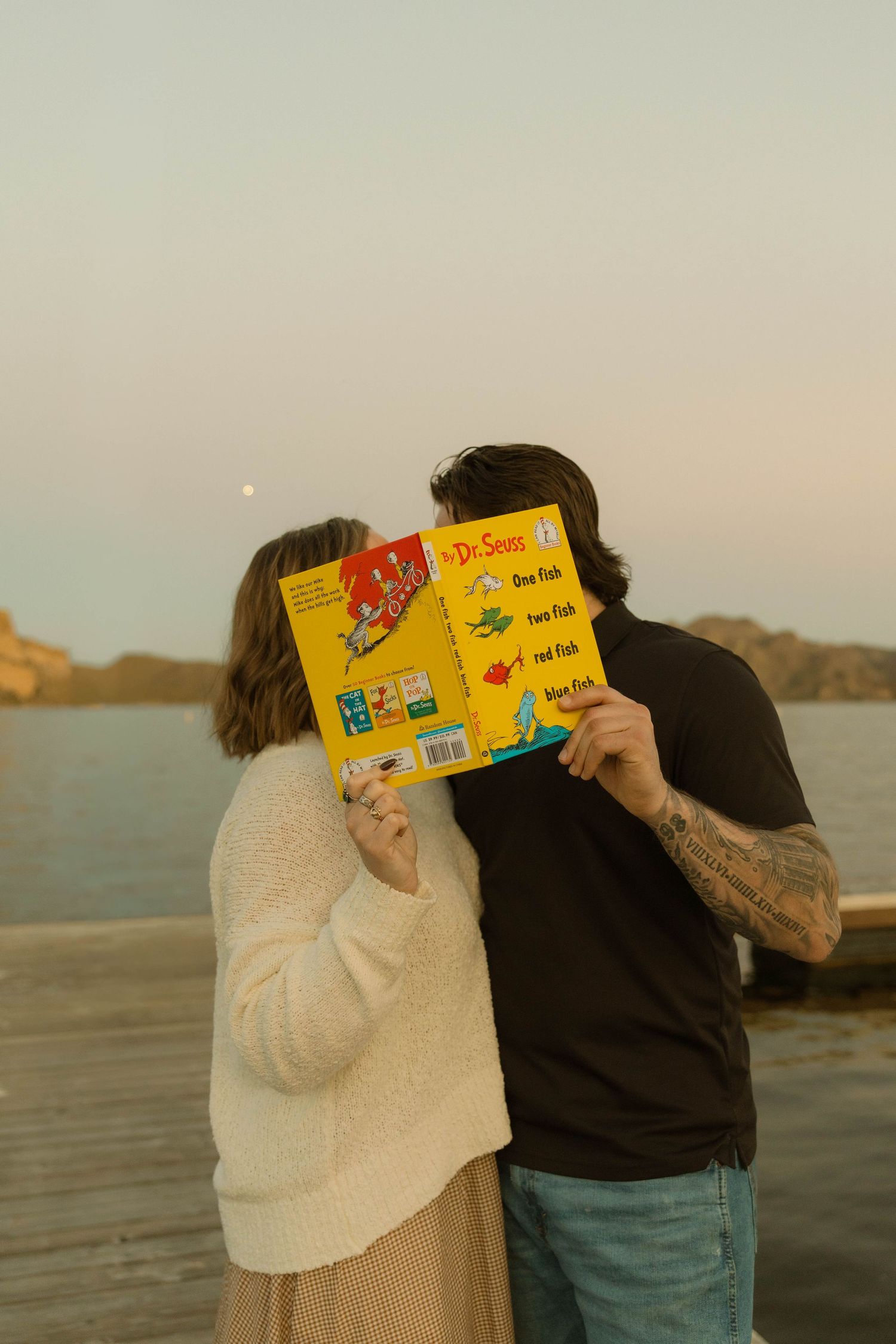 A couple shares an intimate moment behind a colorful children's book by the water at sunset.