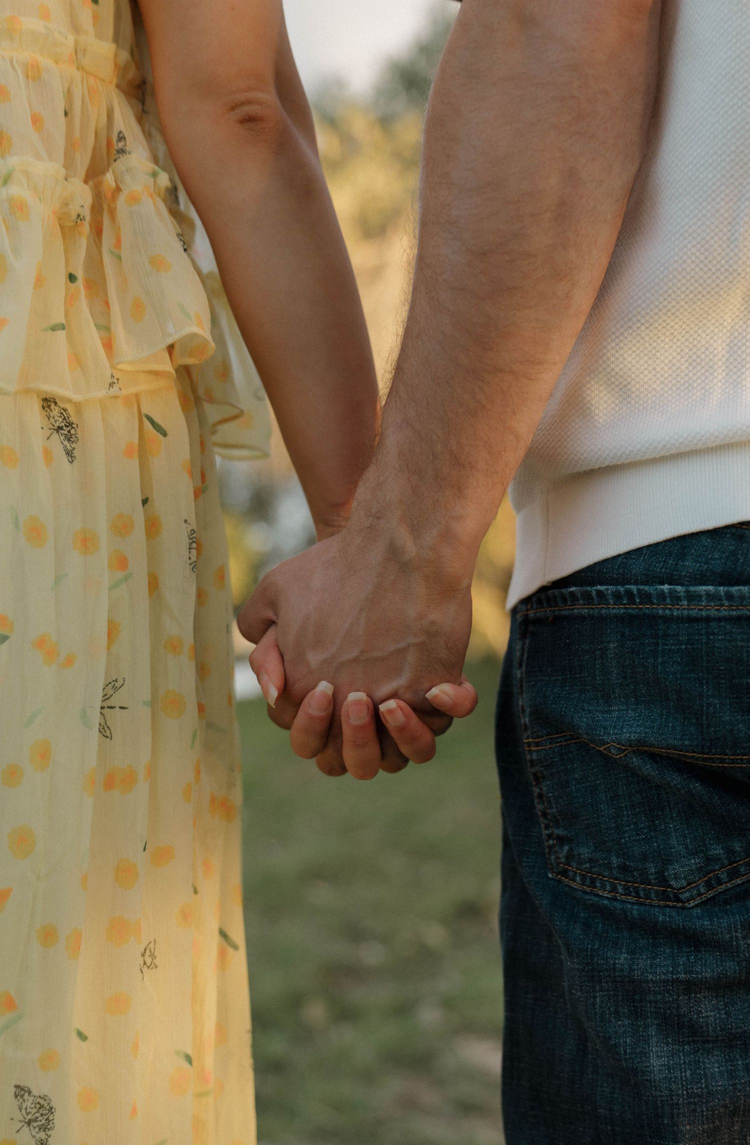 Close-up of a couple holding hands against a yellow floral dress and denim jeans in warm sunlight.