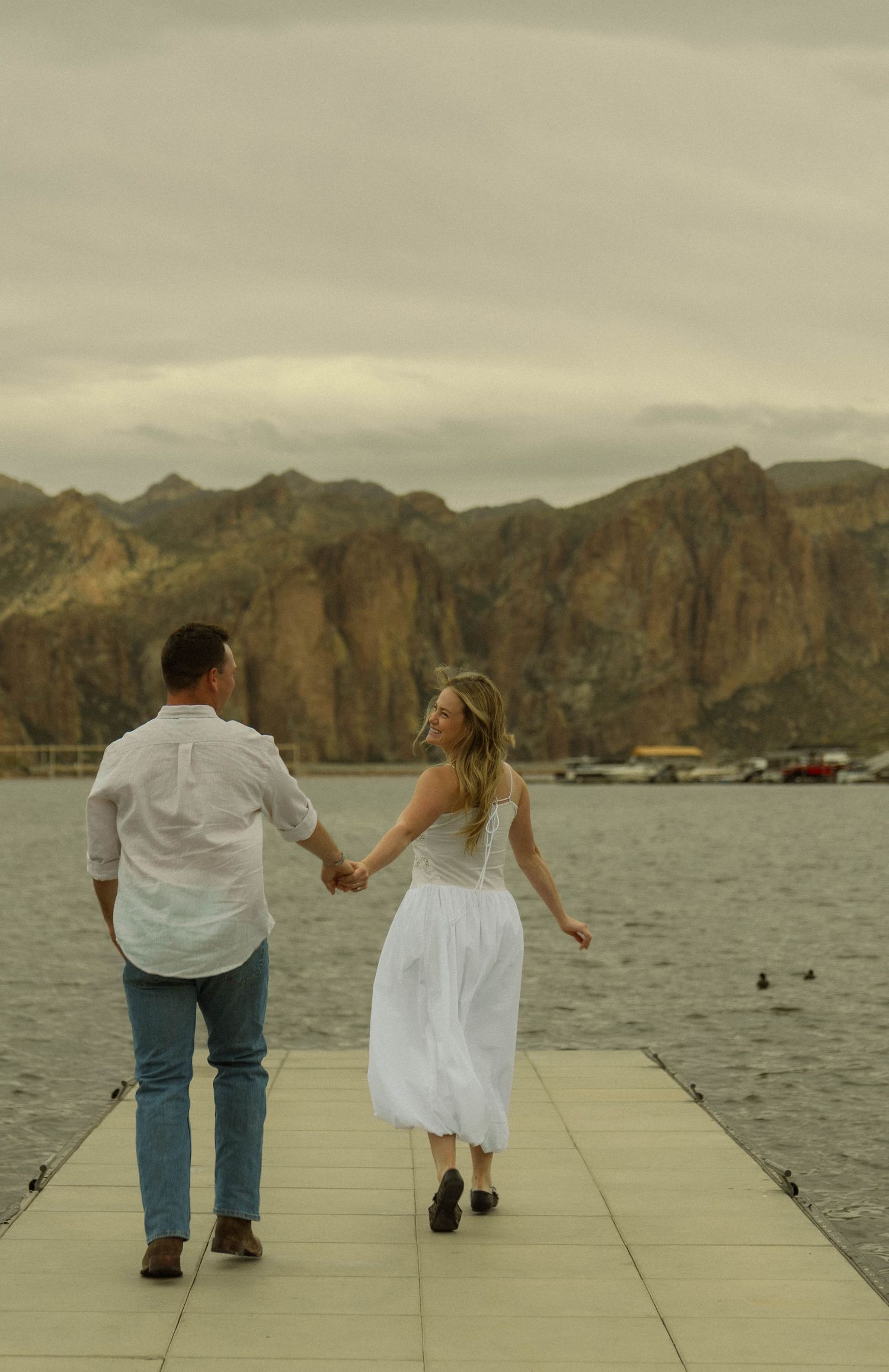 A couple holds hands while walking away on a white dock with mountains and water in view.