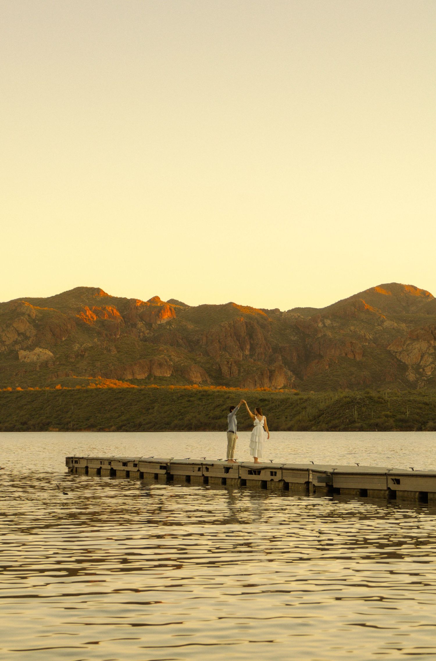 Two people stand at the end of a wooden dock overlooking a serene lake with mountains in the golden sunset light.