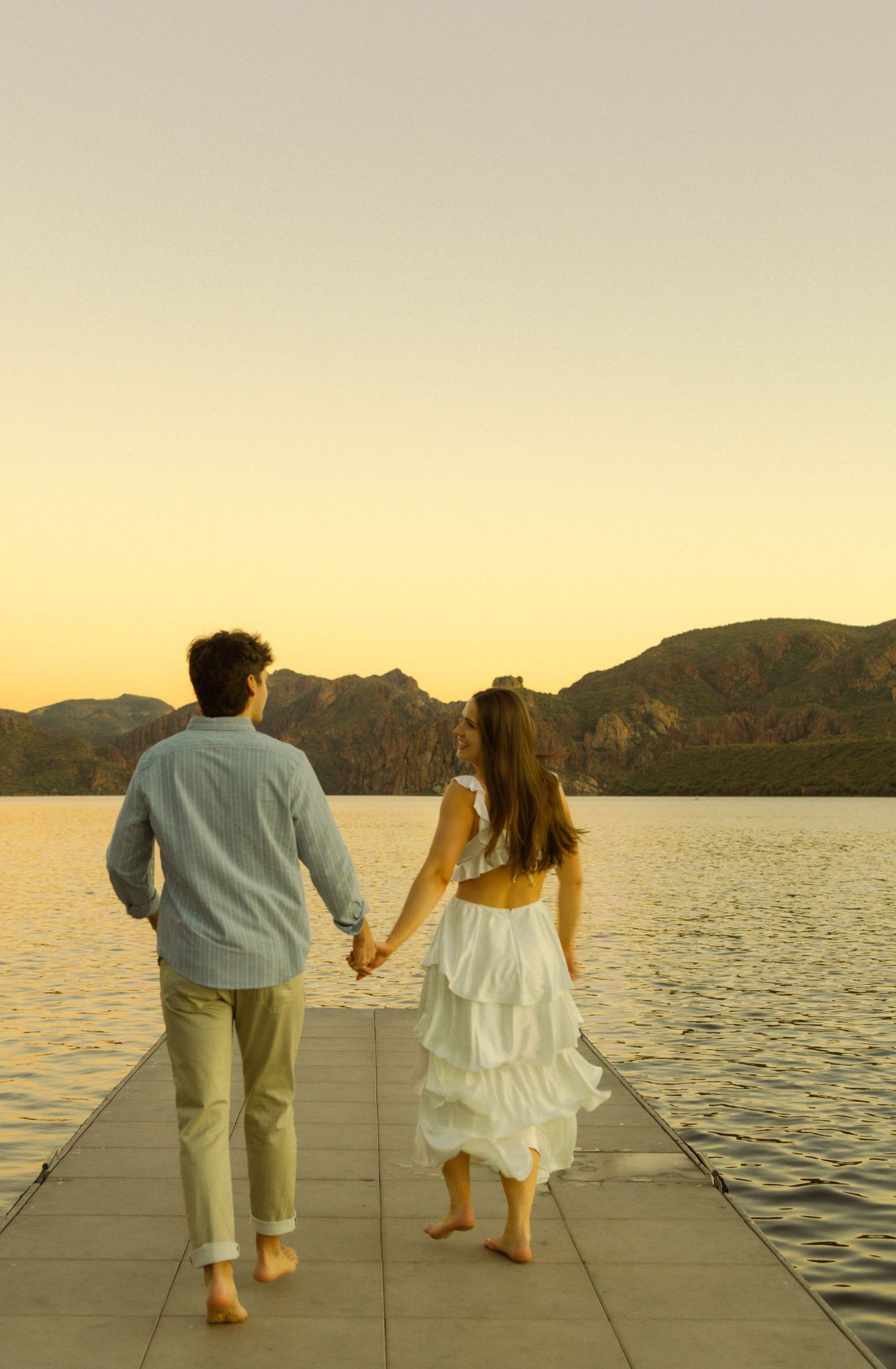 Couple walking barefoot on dock at sunset with mountains and lake in background.