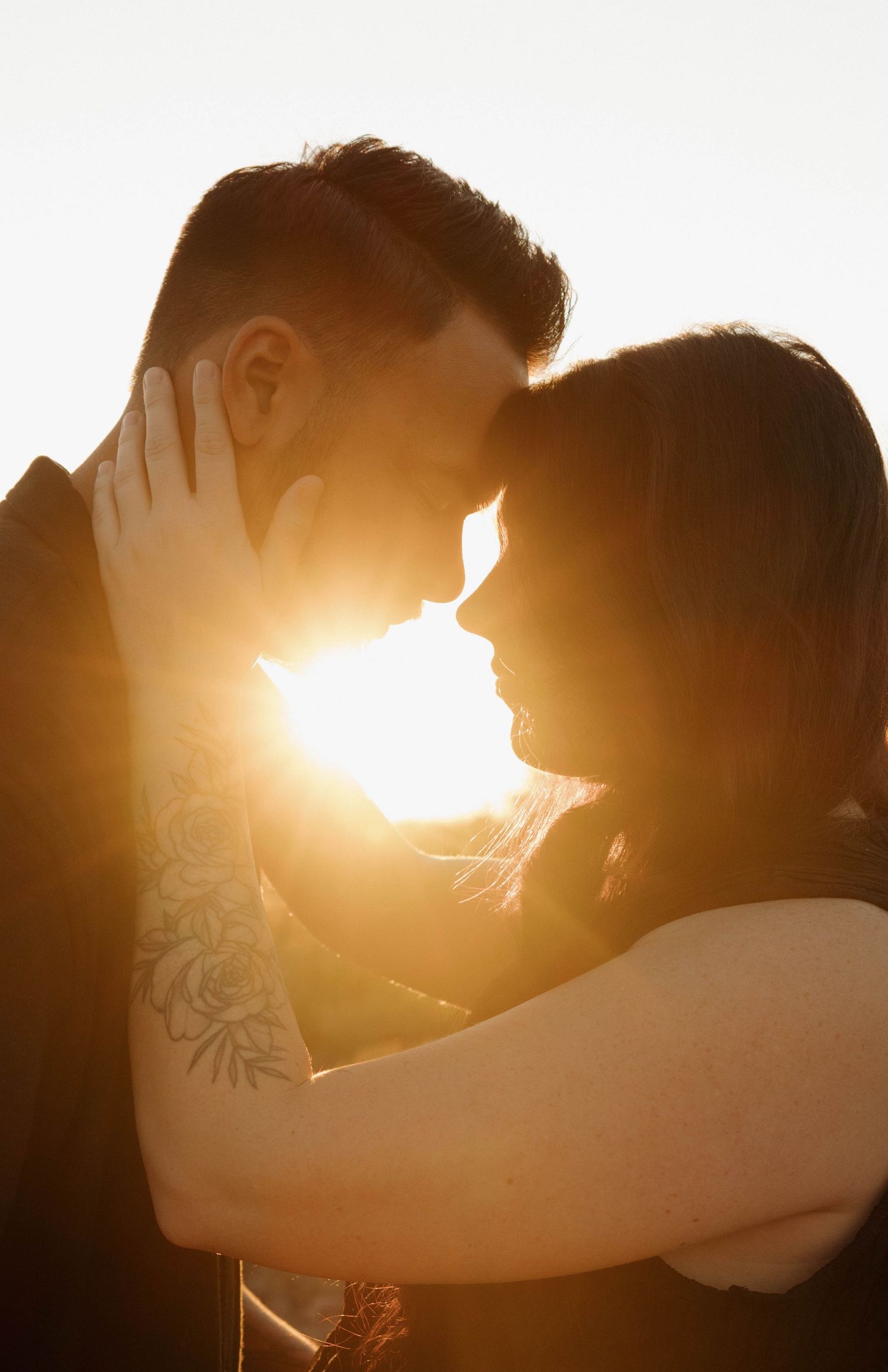 Silhouetted couple embracing in warm sunlight with sun flare between them as one person shows a visible tattoo on their arm.
