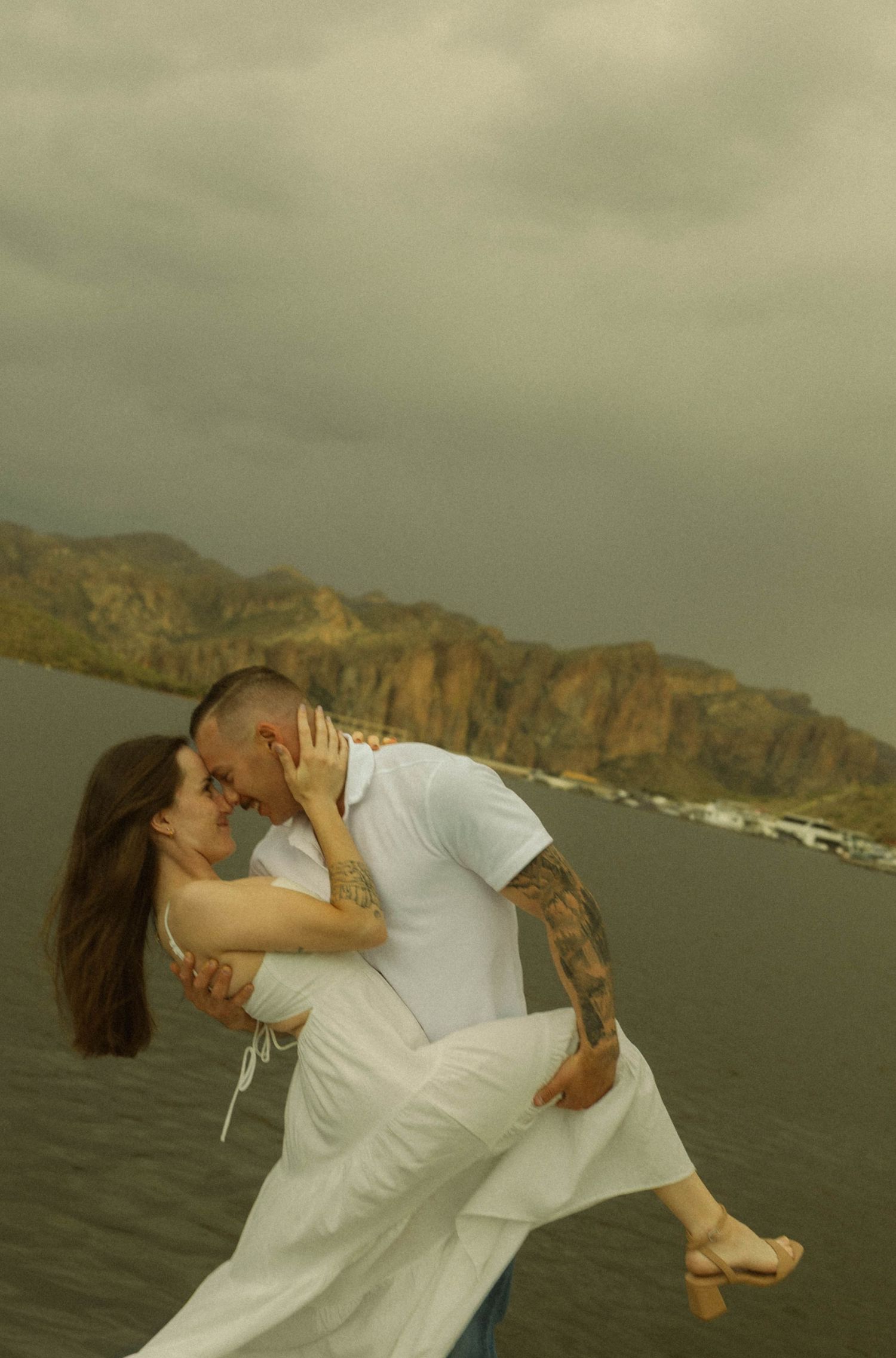 A romantic dip pose captured against dramatic mountain cliffs by the water.