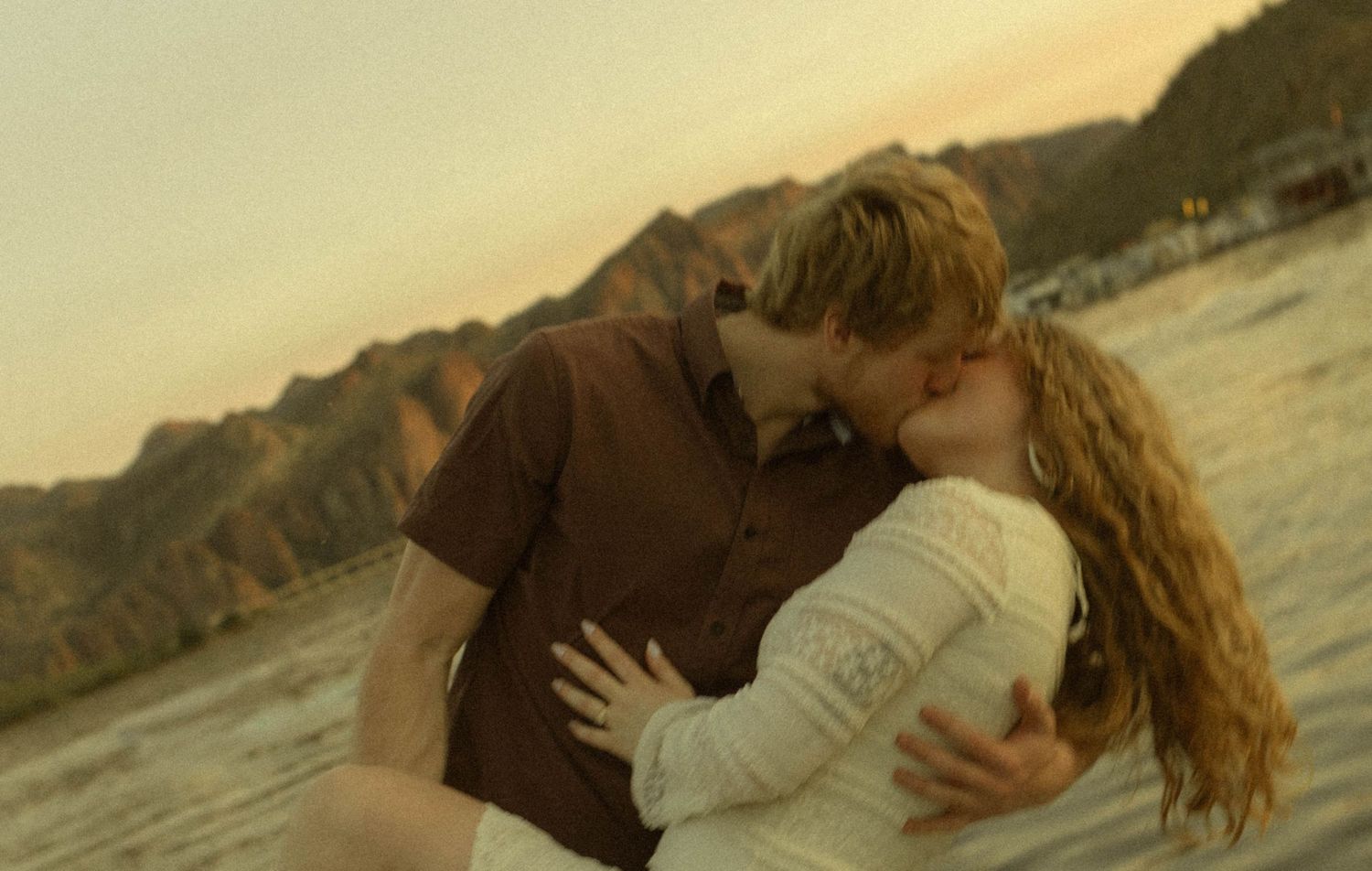 A romantic couple embraces passionately on a beach at sunset with rocky cliffs in the background.