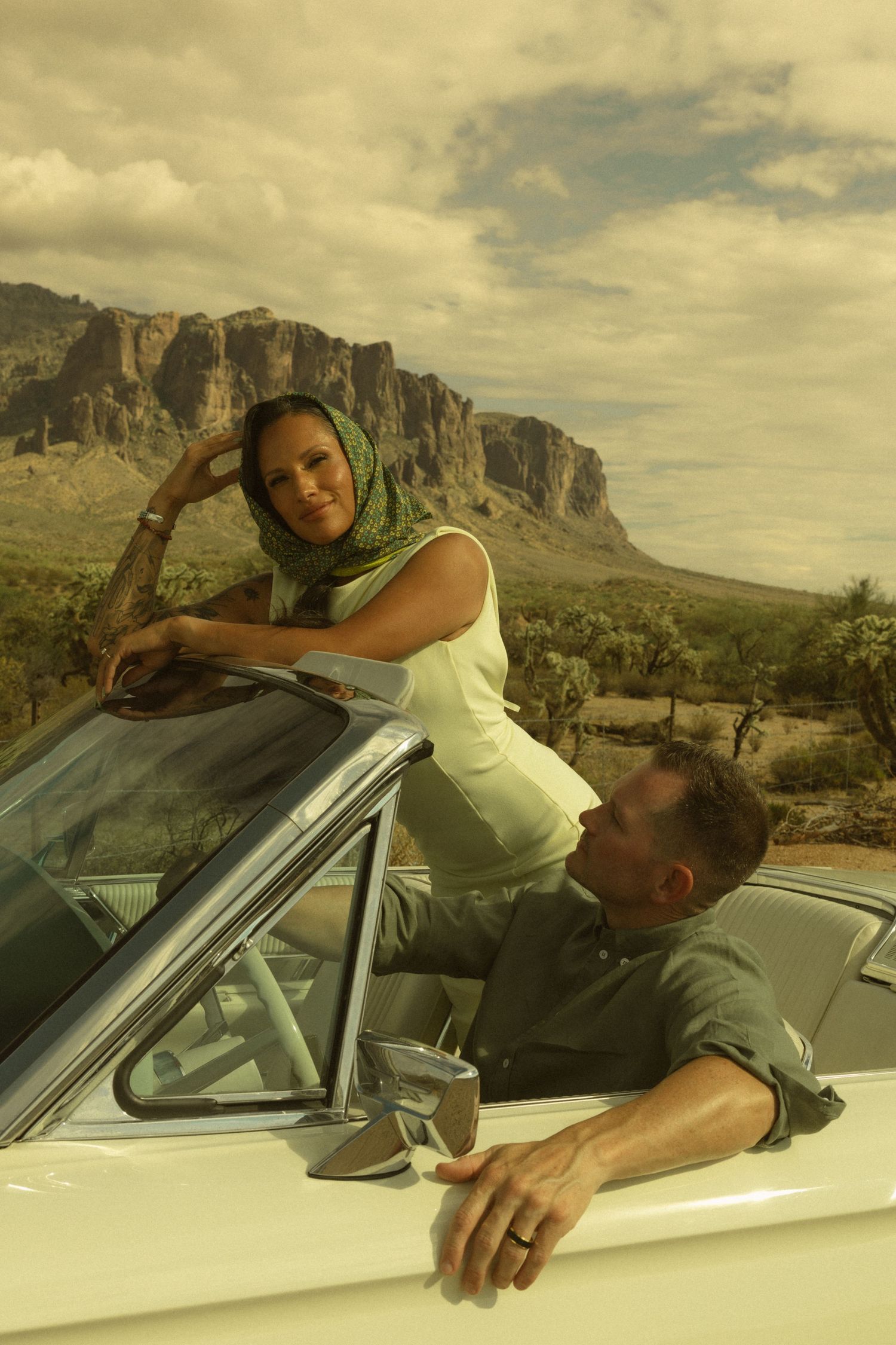 A woman in a green headscarf leans on a vintage white convertible as a man looks up at her in the Arizona desert.