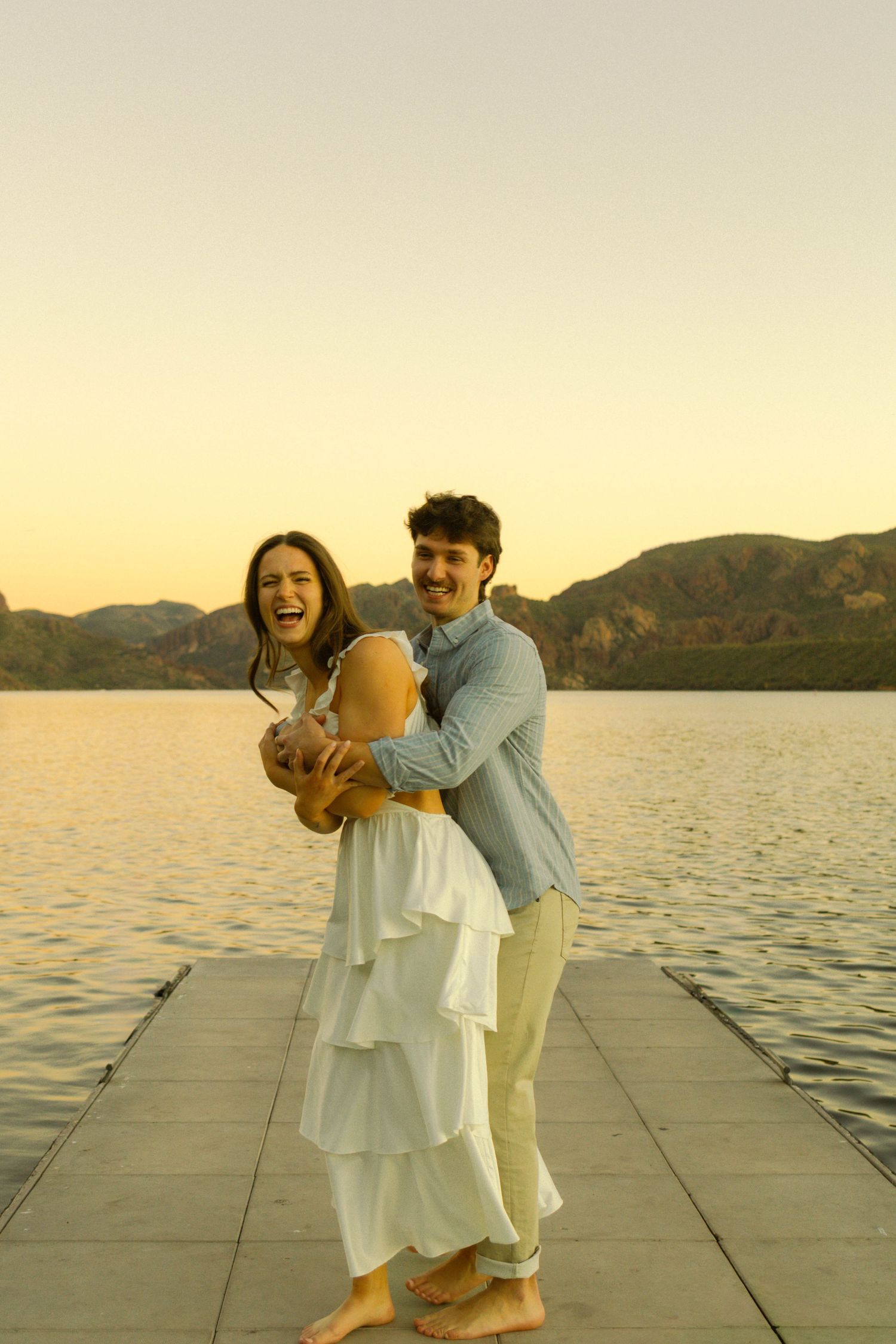 Couple embraces playfully on a wooden dock at sunset with mountains and lake in background.