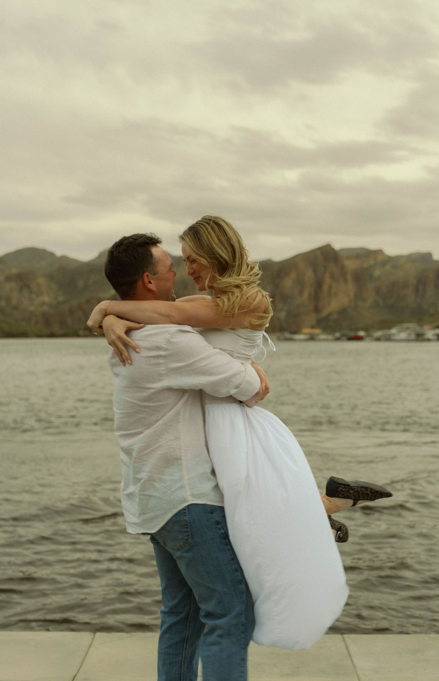 A romantic couple embraces on a boat dock with mountains and water in the background at sunset.