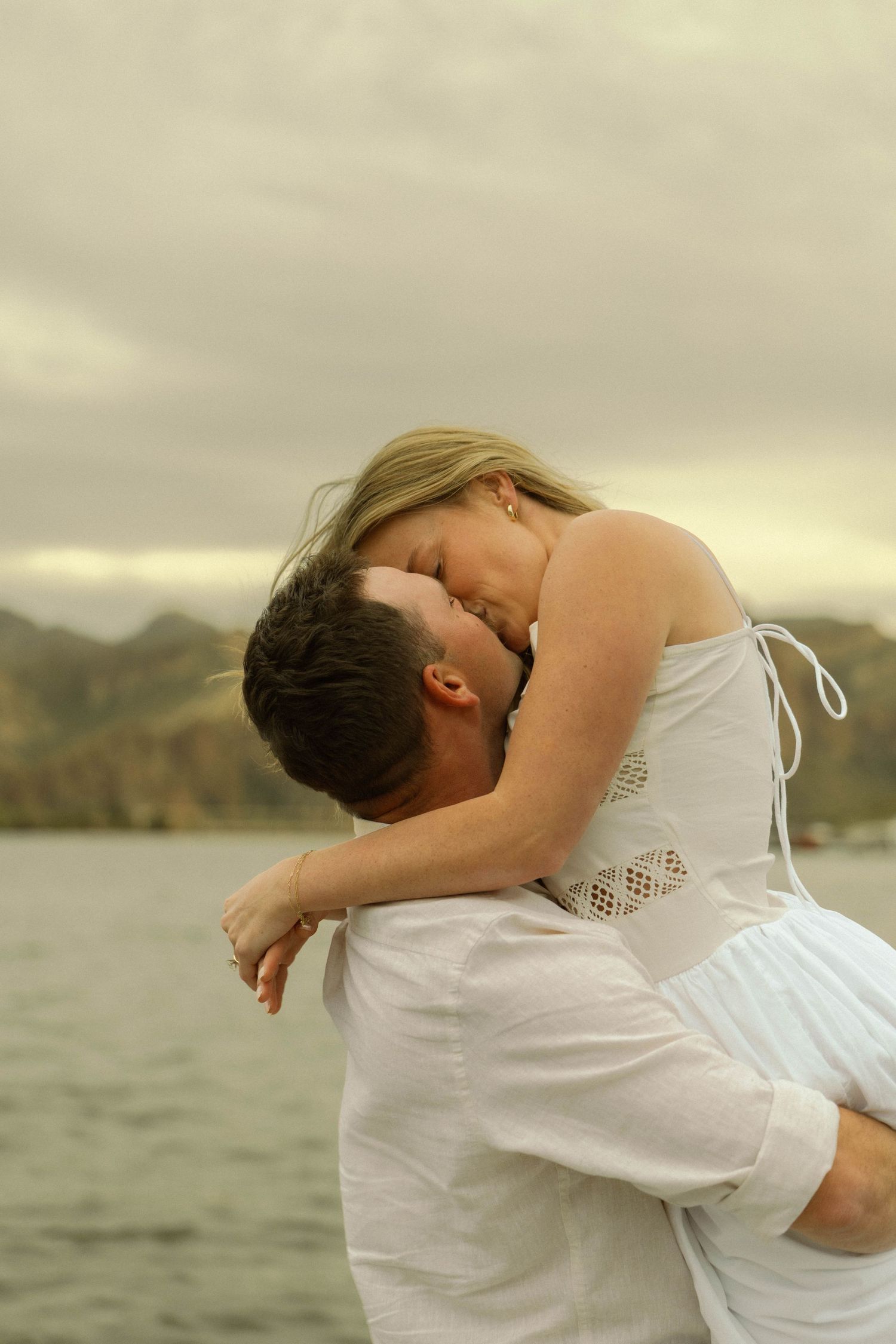 A couple in white clothing embraces playfully by a scenic mountain lake during golden hour.