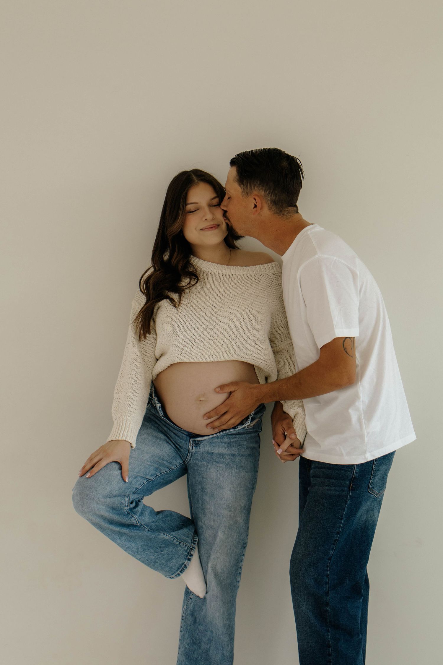 A couple shares an intimate moment during a maternity photoshoot against a neutral wall background.