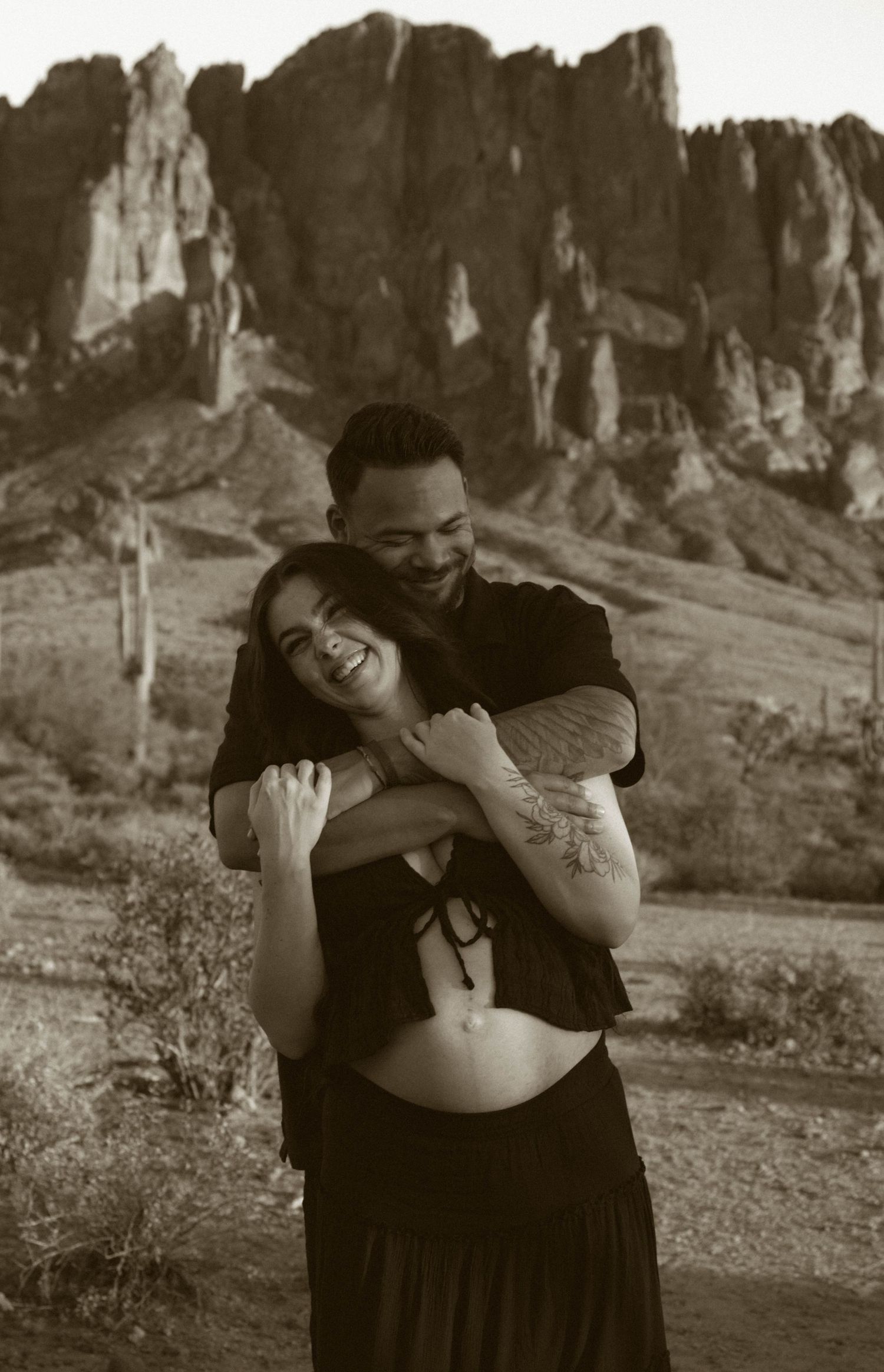 A romantic couple embraces against a backdrop of dramatic desert cliffs and mountains in a black and white photo.