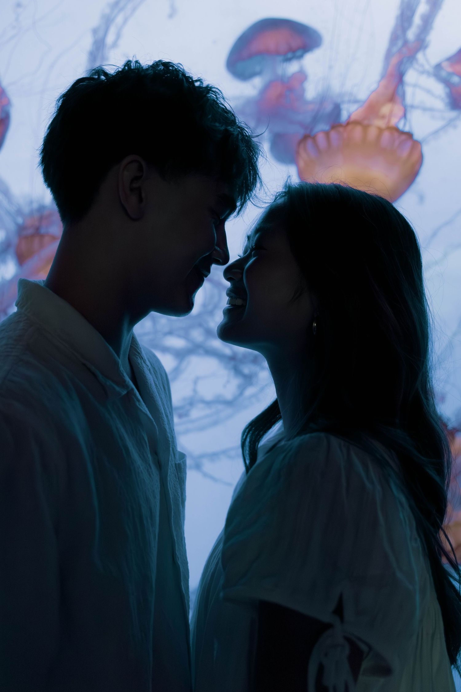 Dark silhouettes viewed through aquarium glass with floating jellyfish in the background.
