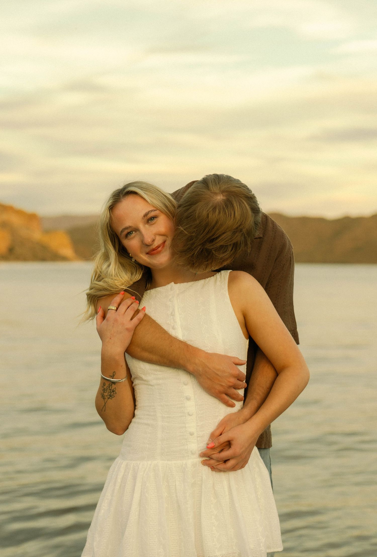 Couple shares playful moments on a lakeside at golden hour with mountains visible in the distance.
