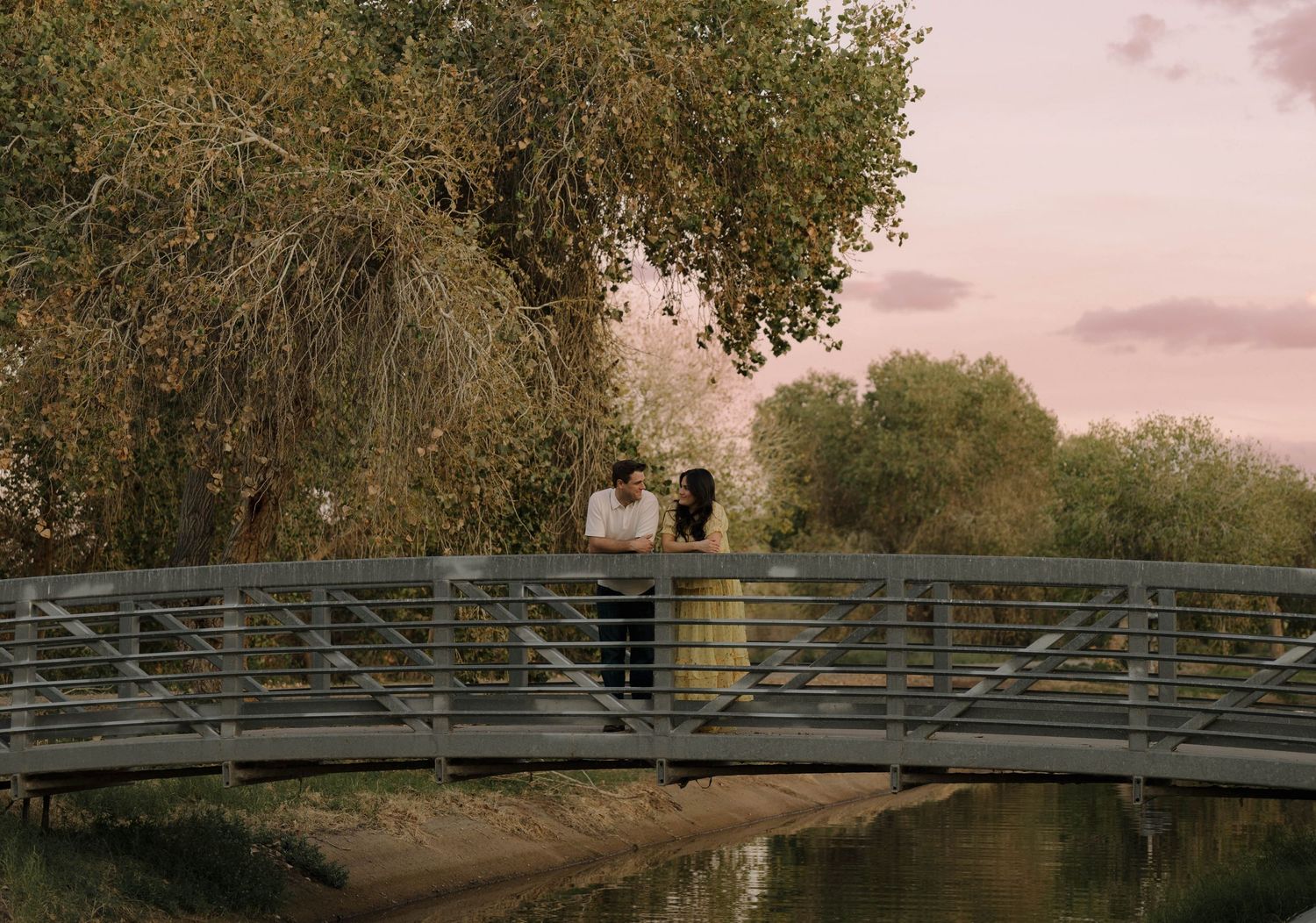 Row of rustic wooden bridges spanning across a tranquil river at sunset with trees in the background.