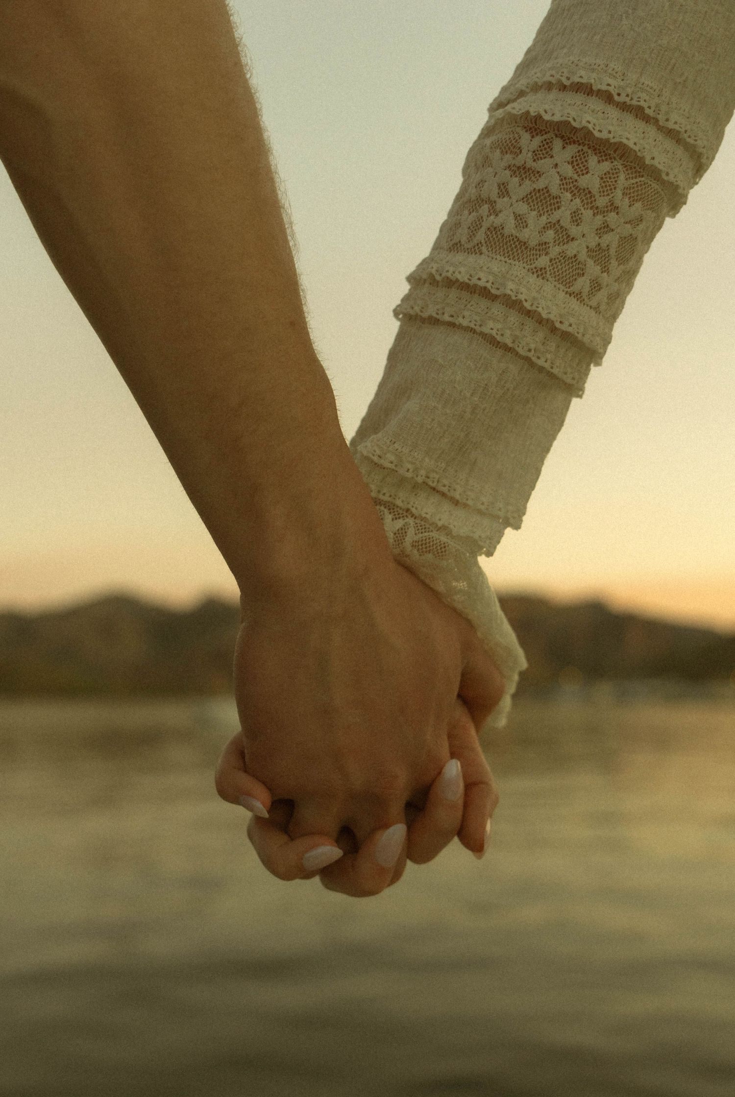 Two people holding hands intimately at sunset by a lake, with one wearing a decorative knit sleeve.