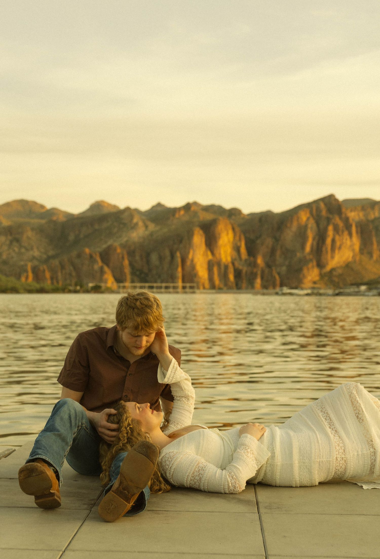 Couple lying on a dock by a scenic lake with mountainous cliffs at sunset in warm golden light.
