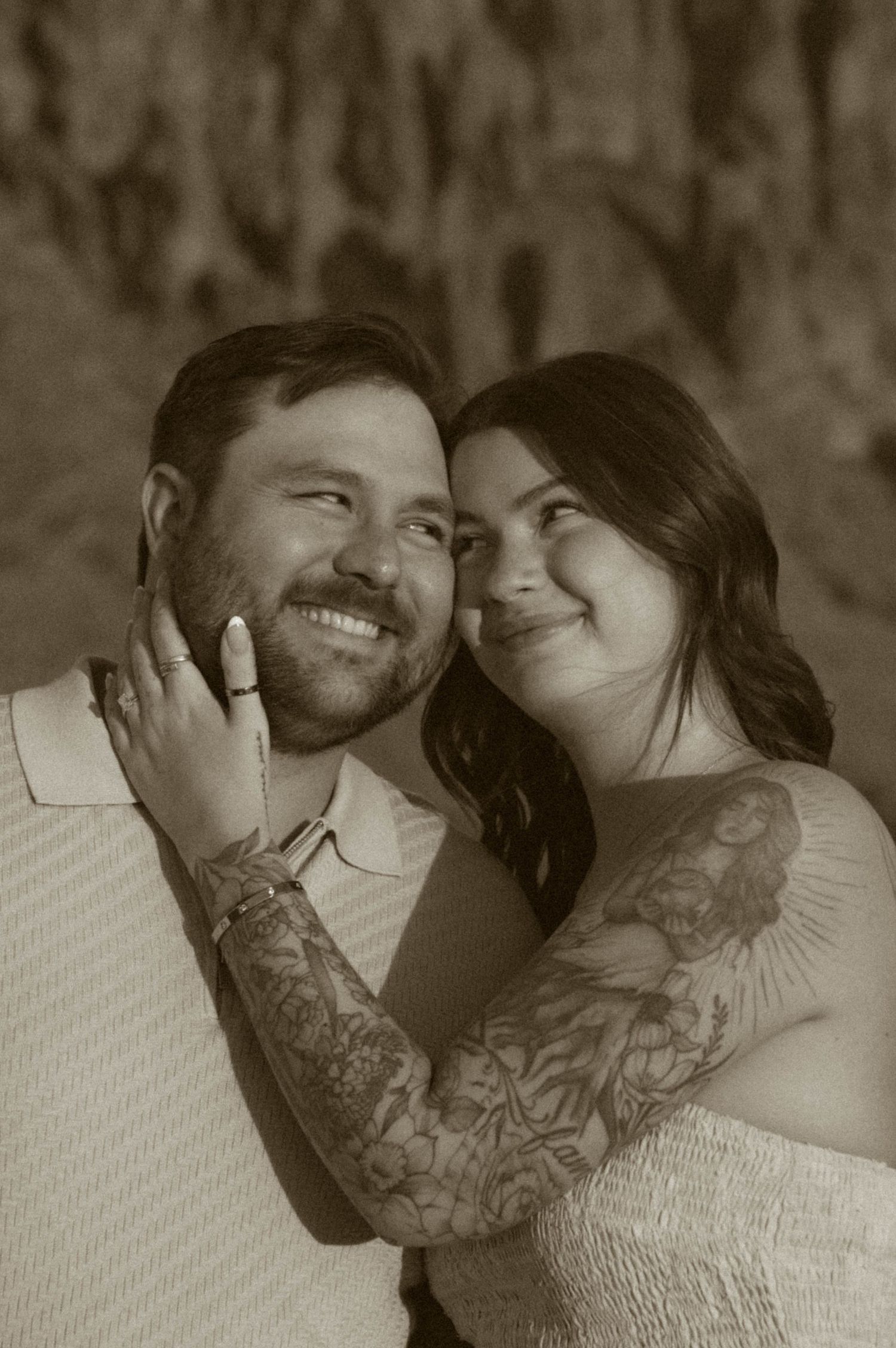 A smiling couple with tattoos poses intimately in a sepia-toned engagement photo, the woman tenderly holding the mans face.