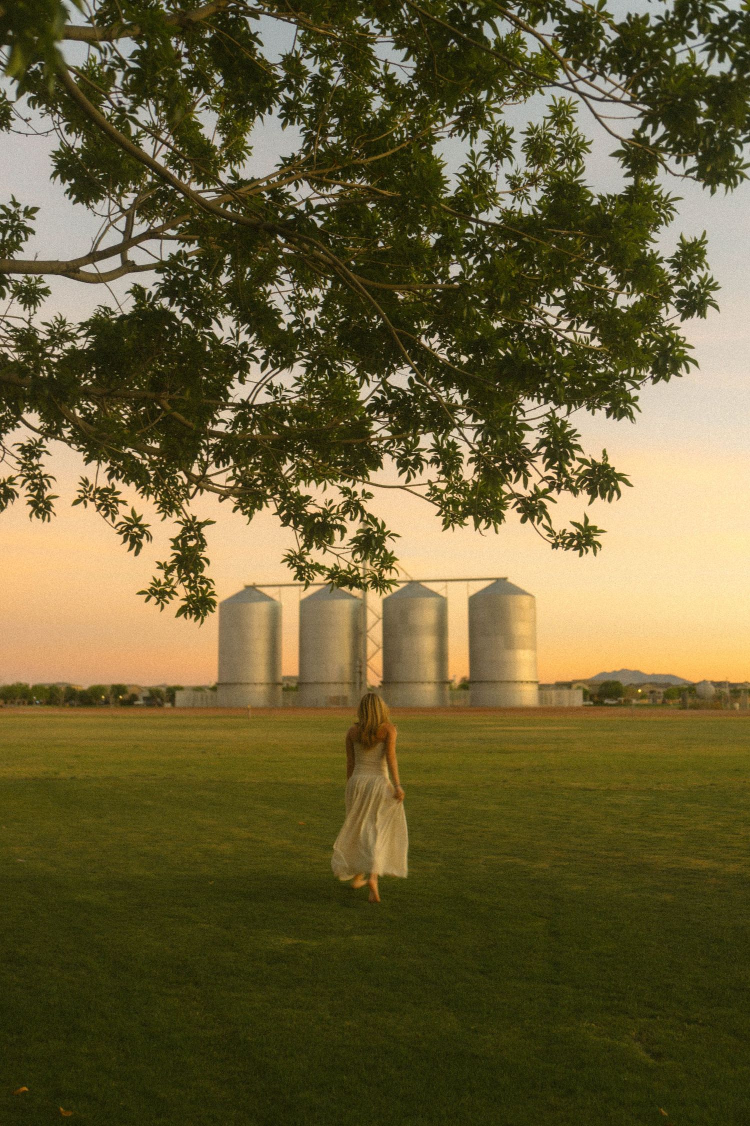 Blonde woman in white dress walking toward grain silos under a glowing sunset sky.