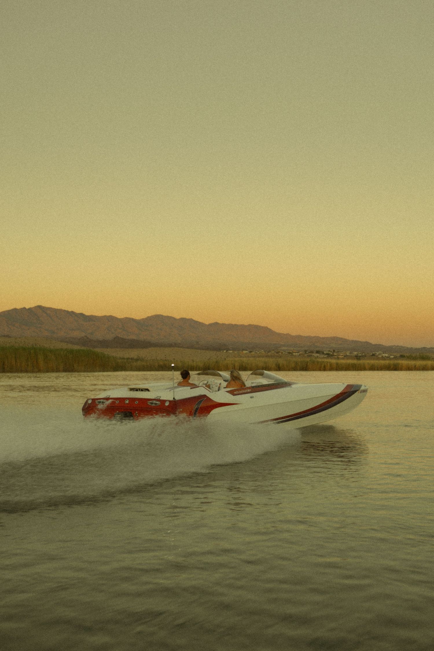 Red and white speedboat racing across a lake at golden sunset with mountain silhouettes.