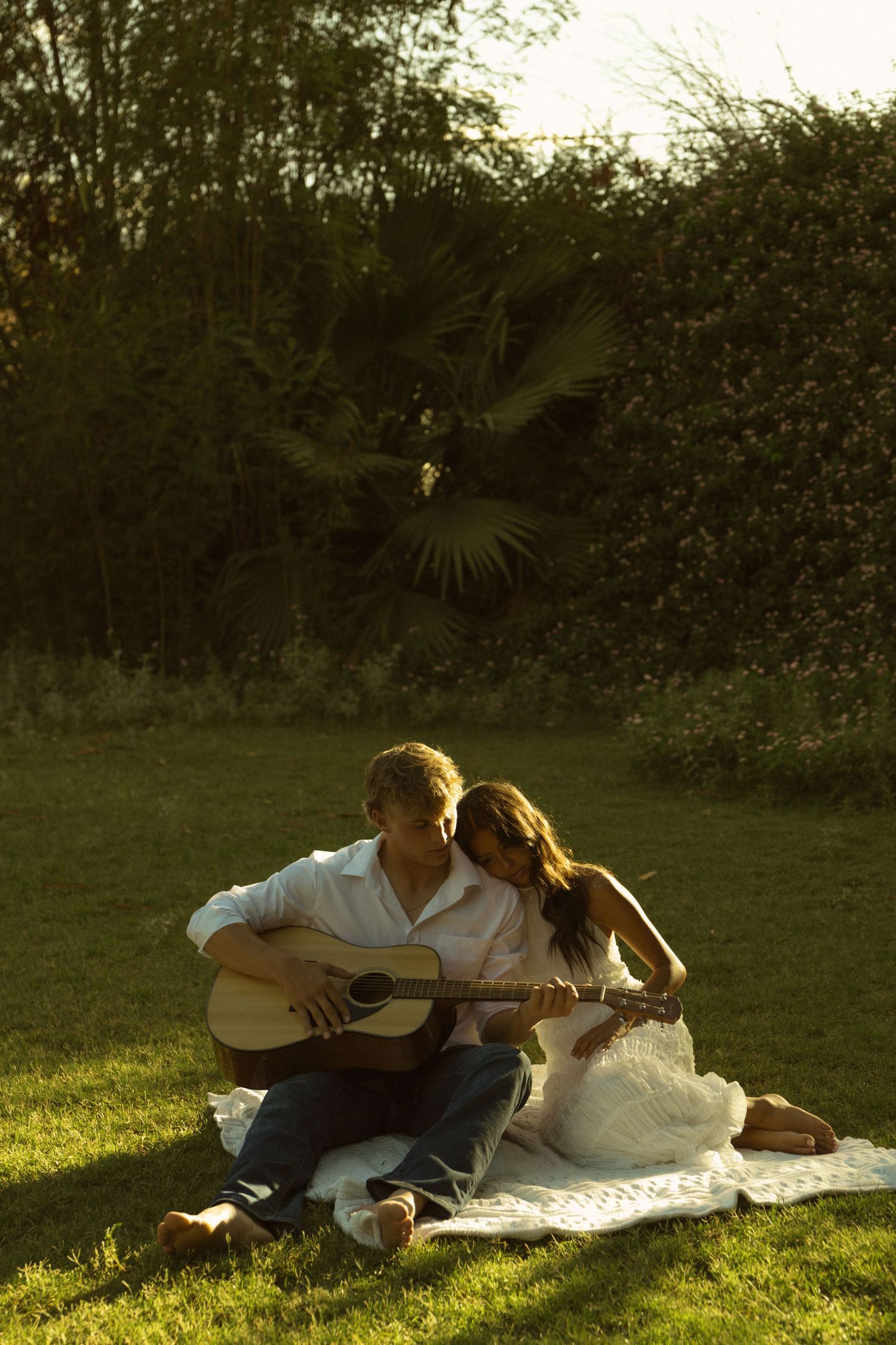 Couple sitting on blanket in garden at golden hour, man playing acoustic guitar while woman leans on his shoulder.