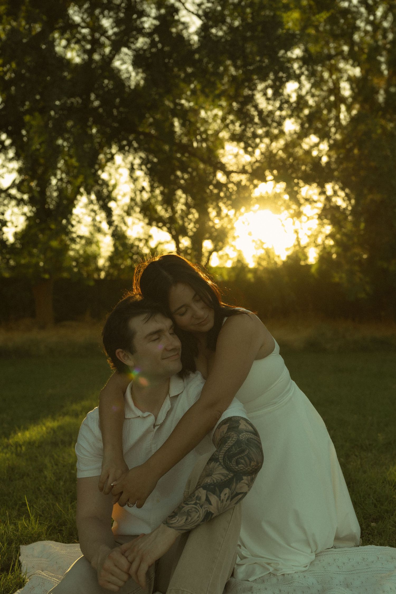 Couple embracing at golden hour sunset in a park, woman in white dress with tattooed arm hugging man from behind.