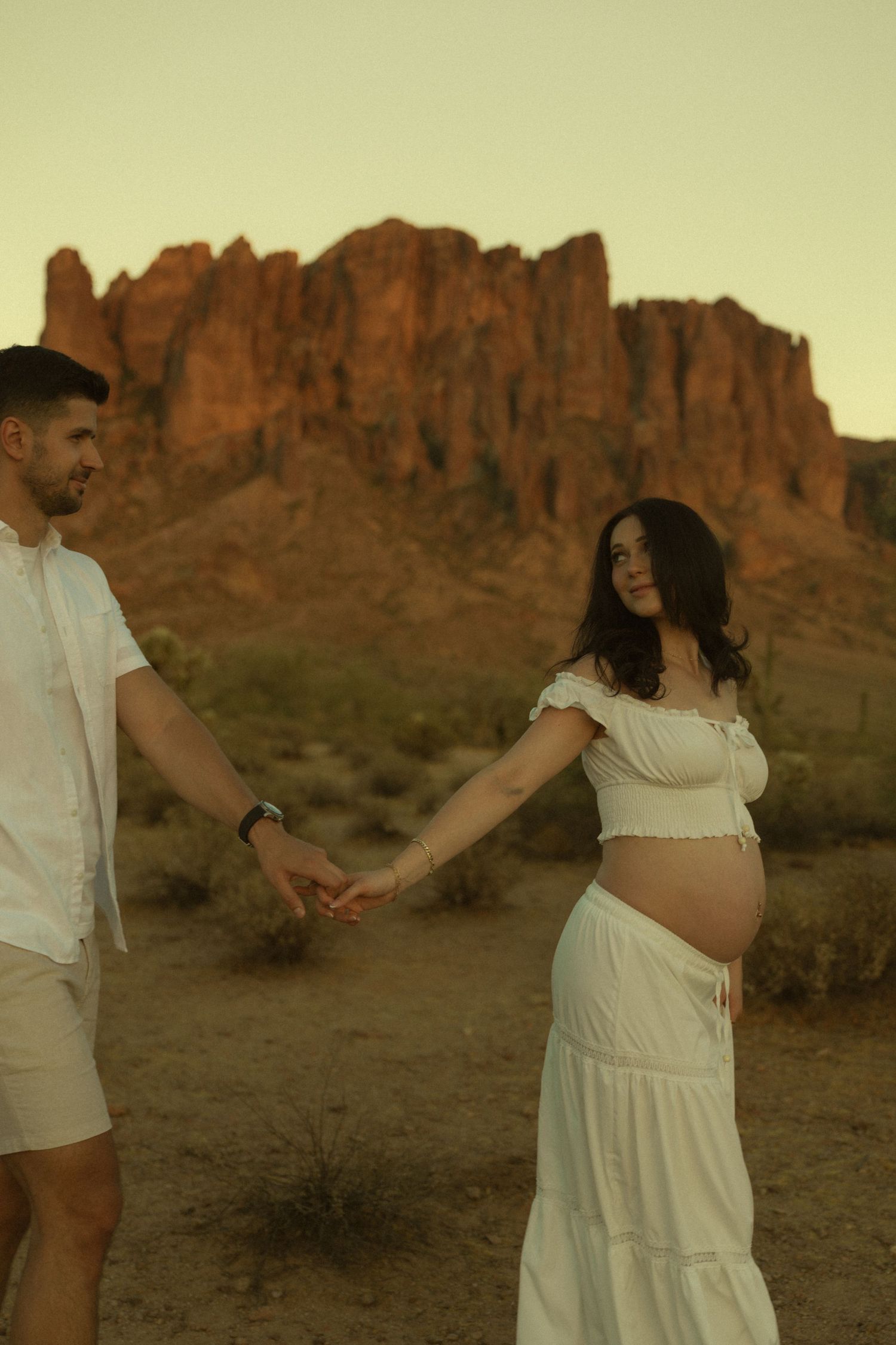 Pregnant couple holding hands in desert at golden hour with Superstition Mountains backdrop in Arizona.