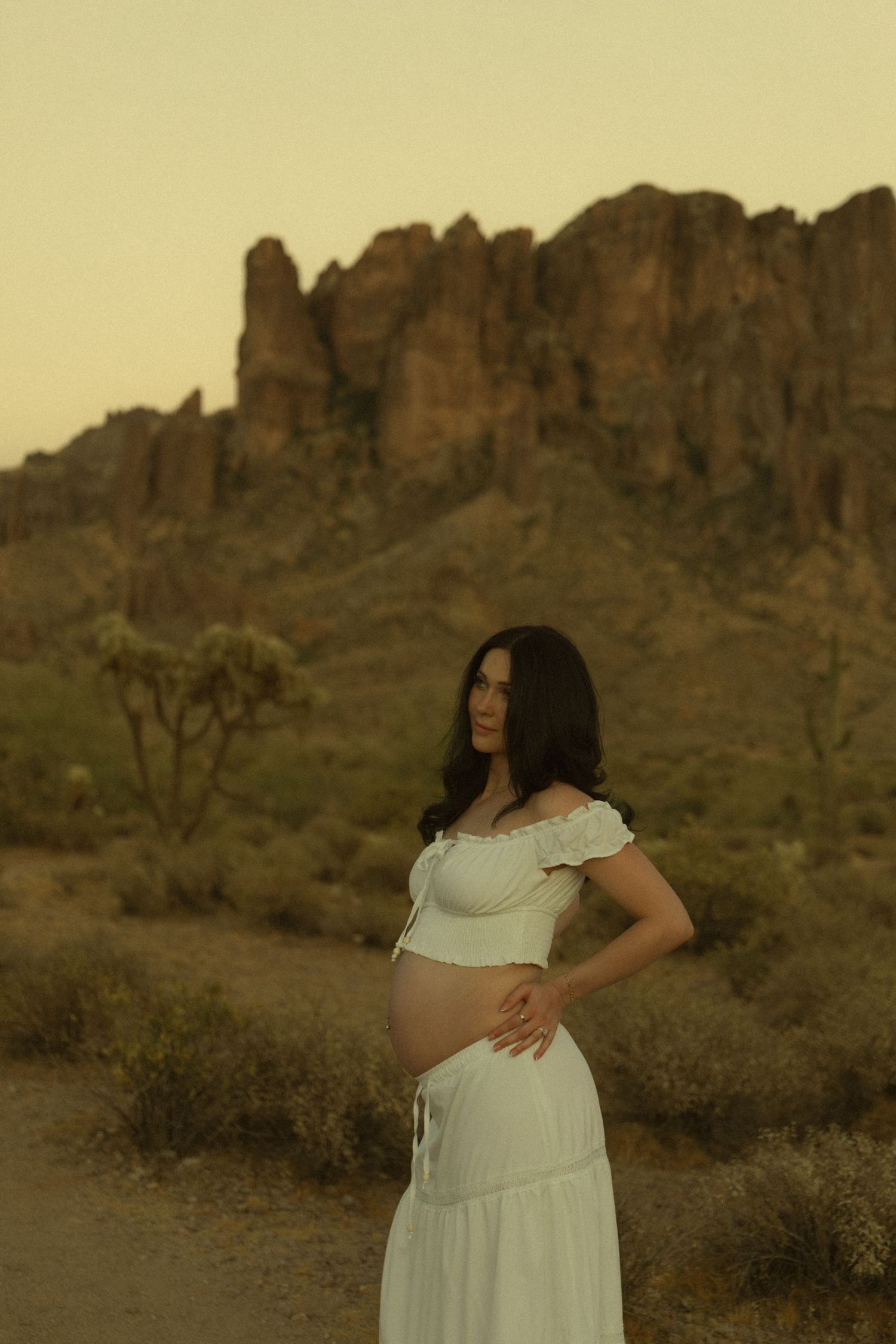 Pregnant woman in white crop top and skirt posing before Superstition Mountains at desert sunset.