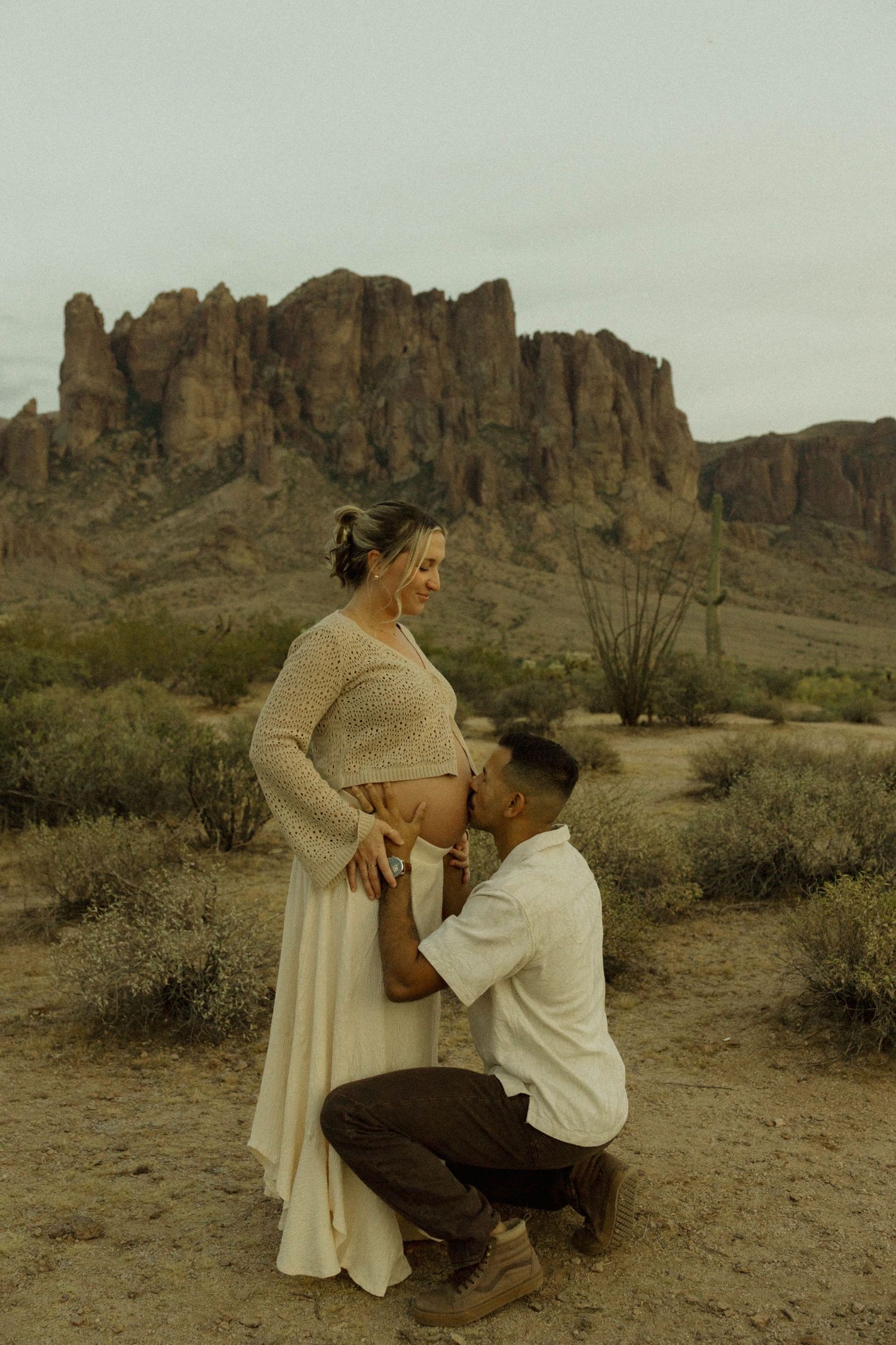 Couple shares maternity photo in Arizona desert, man kneeling kissing pregnant belly with rocky mountain backdrop.