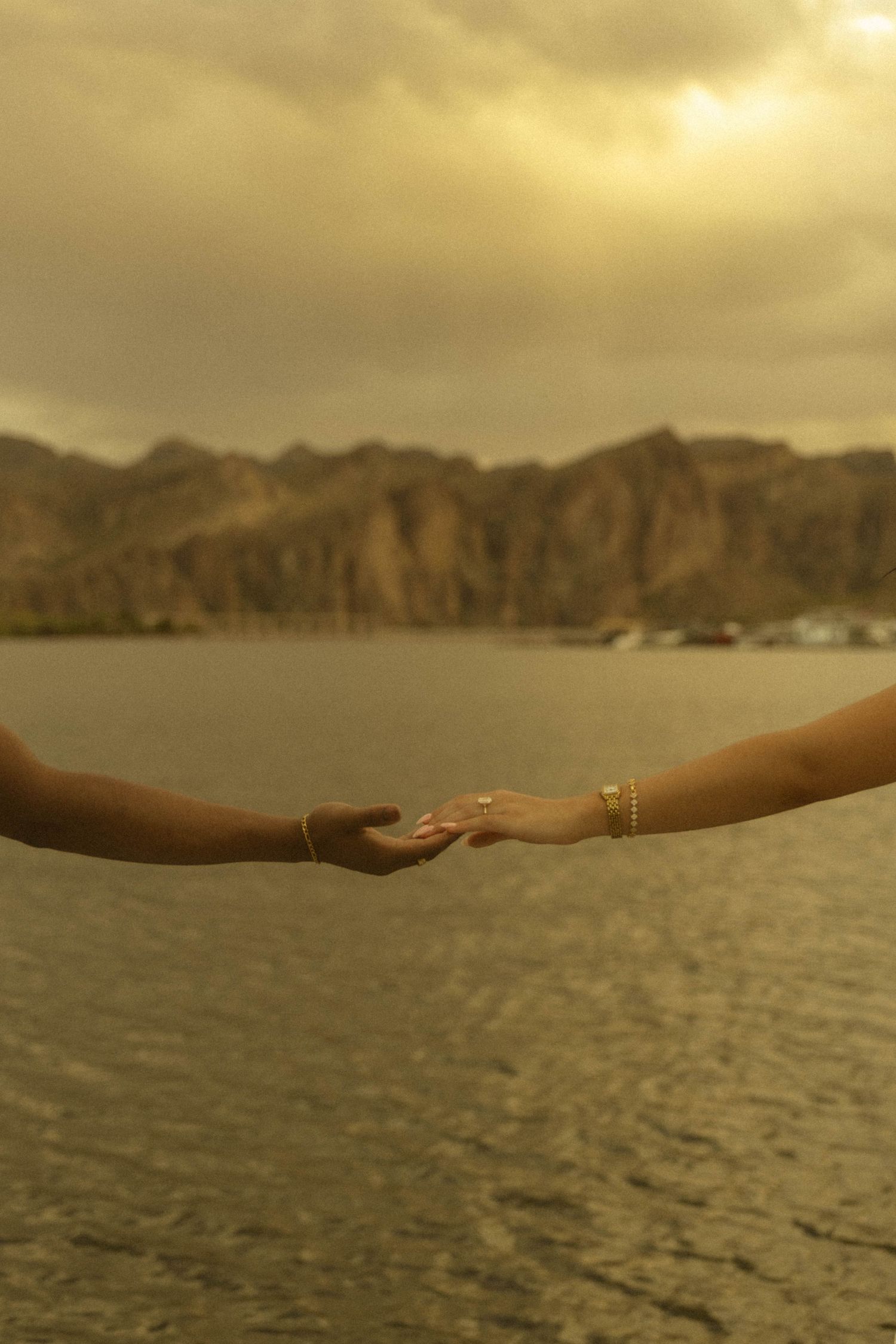 Two hands reaching toward each other with a golden lake and mountain backdrop at sunset.