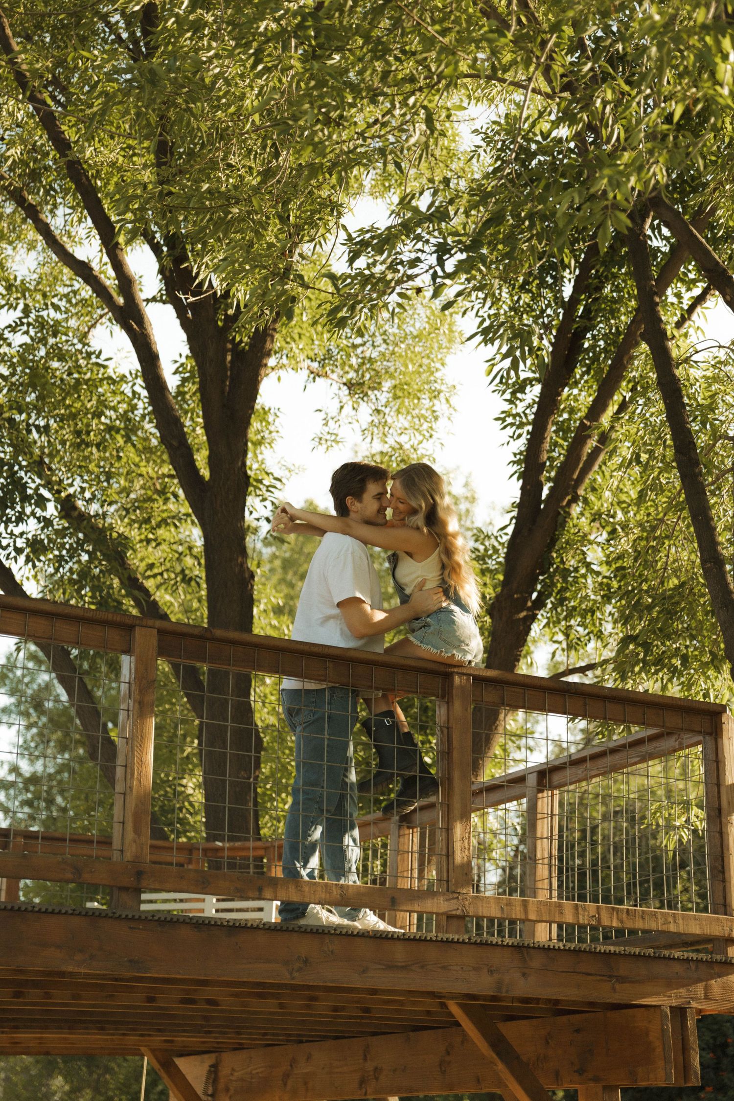 Young couple embracing and kissing on an elevated wooden deck surrounded by green trees.