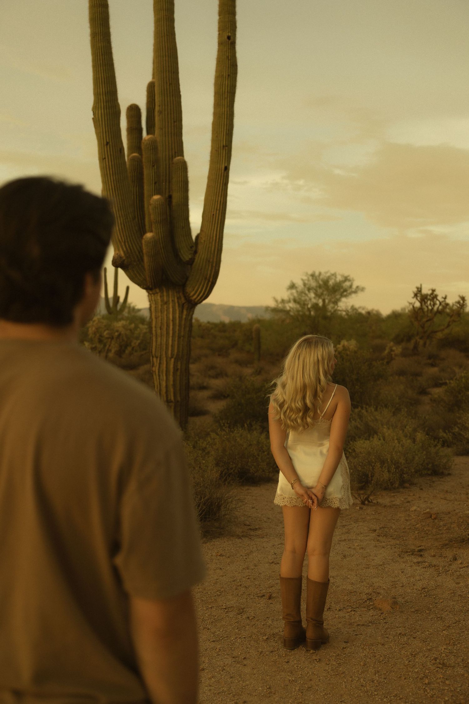 Blonde woman in white dress and cowboy boots stands on desert trail near saguaro cactus at sunset.