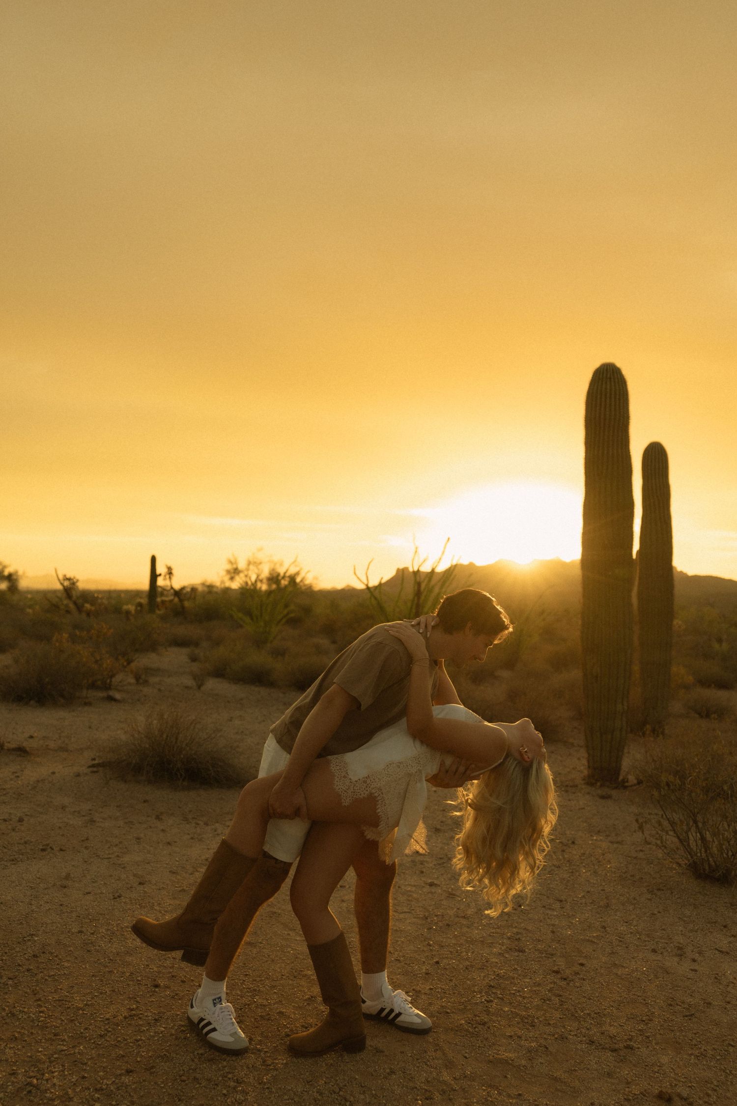 Couple sharing a dip kiss silhouetted against vibrant golden sunset with saguaro cactus.